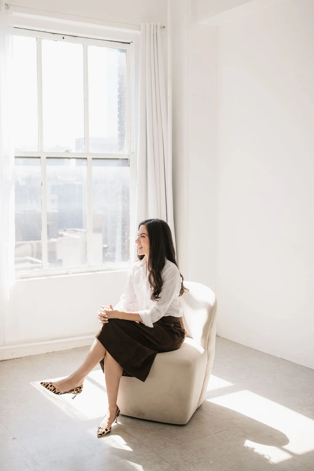 A woman sitting on a white armchair near a large window with white curtains, wearing a white blouse and a dark skirt, with leopard print high heels, smiling and looking to her right.