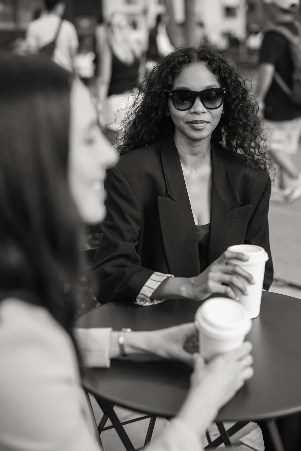 Two women sitting at an outdoor table with coffee cups. The woman in the background is wearing sunglasses and a blazer, facing slightly to the right, smiling. The woman in the foreground is partially visible, with her face blurred, holding a coffee cup.