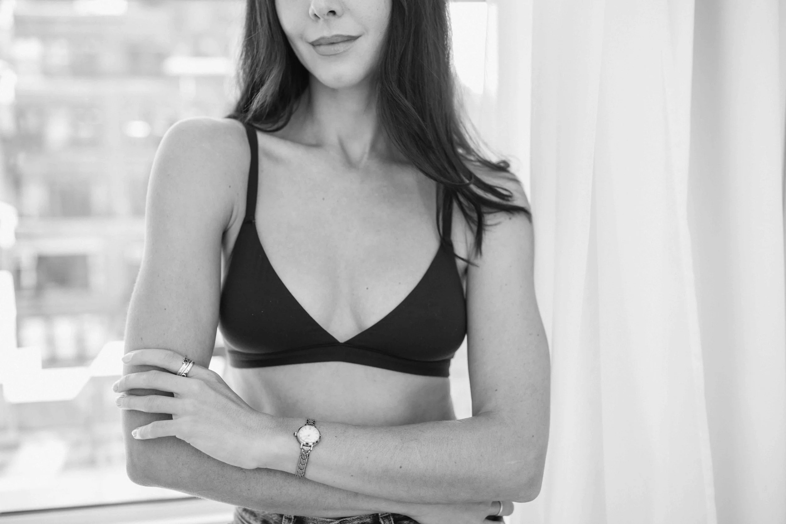 A woman with long hair wearing a black bra, a watch on her left wrist, and a ring on her right hand, posing indoors with a city view in the background.