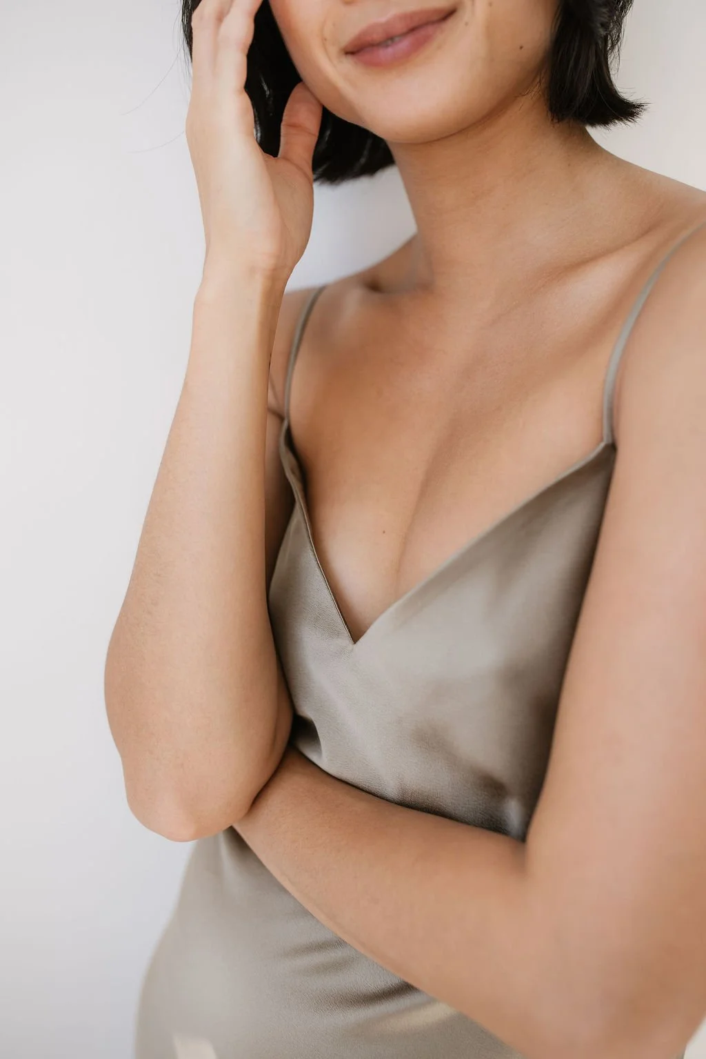 A woman with short black hair wearing a satin beige slip dress, touching her face with her left hand, against a plain white background.
