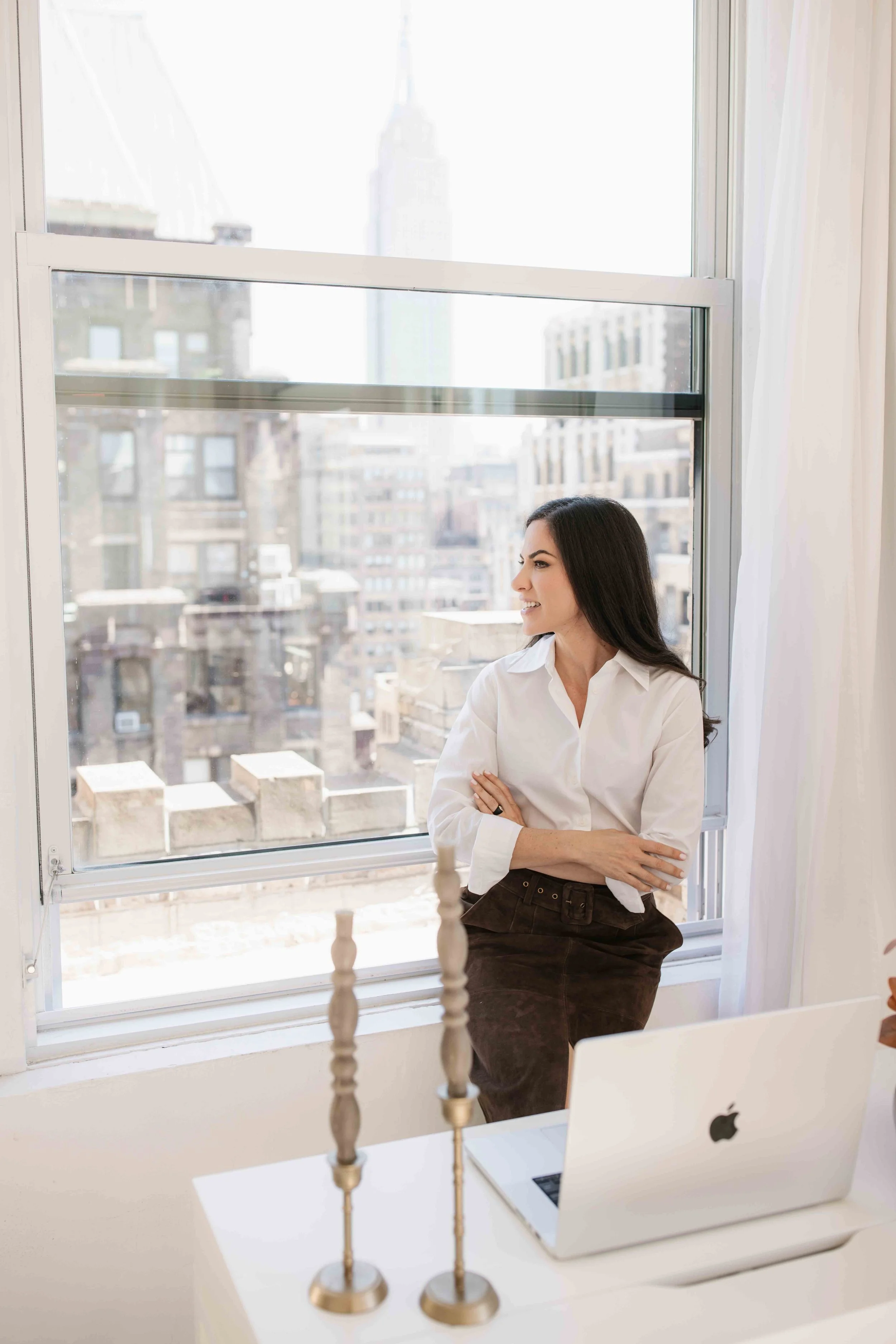 Woman sitting on window sill looking outside in a bright room with city buildings in background.