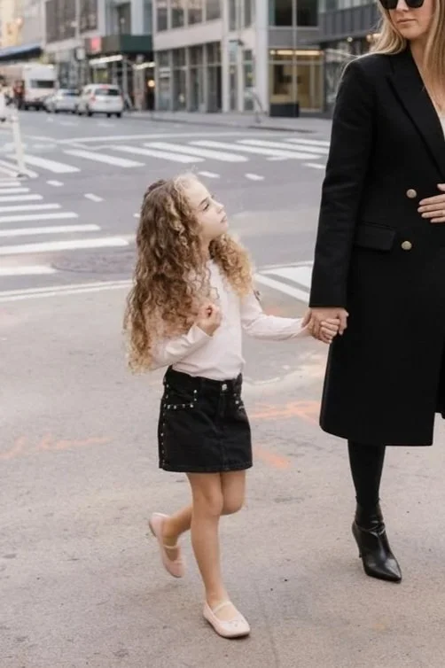 A young girl with long curly blonde hair holding hands with an older woman in a black coat and sunglasses on a city street.