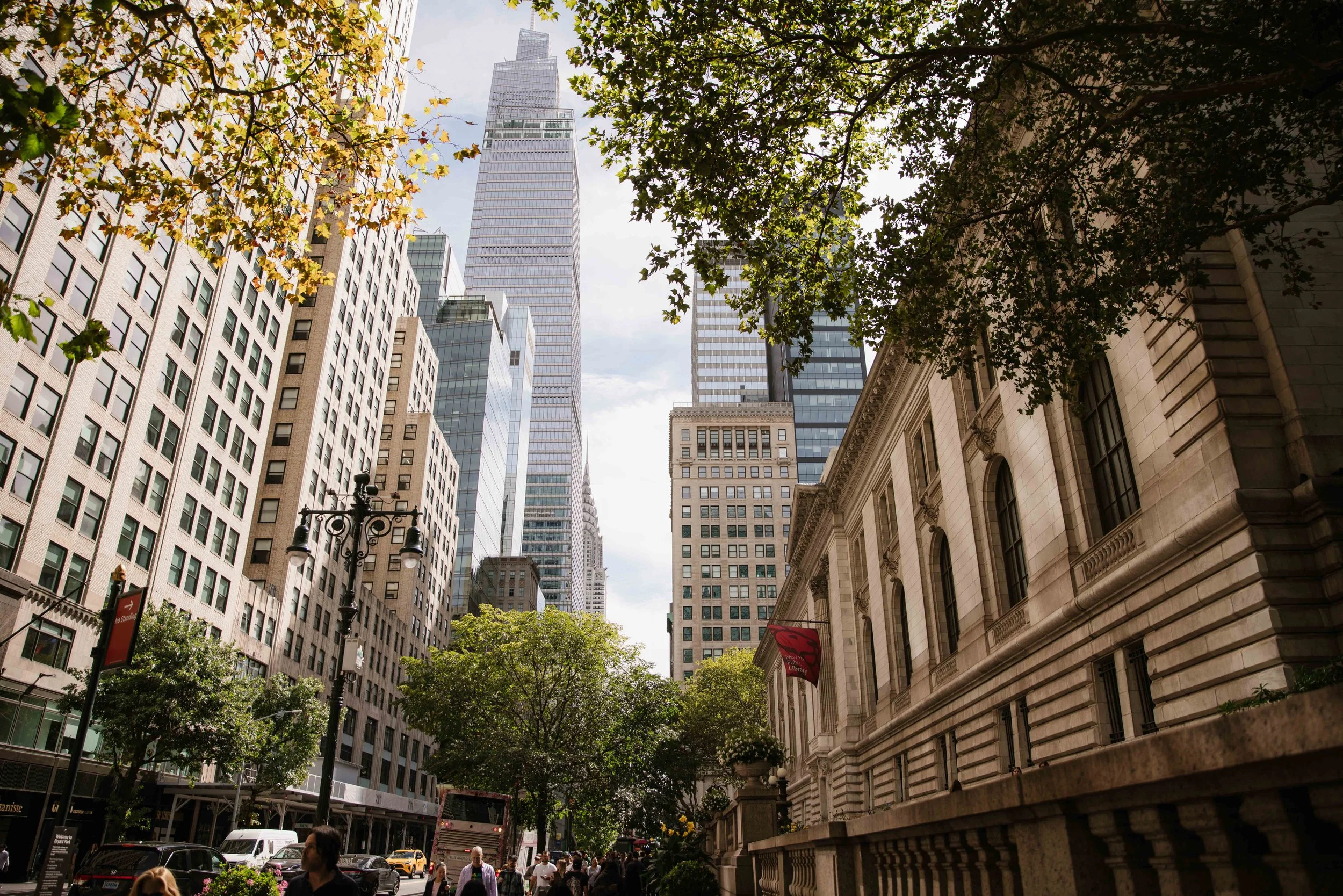 A city street scene with tall skyscrapers and historic buildings, trees lining the sidewalk, and pedestrians walking along.