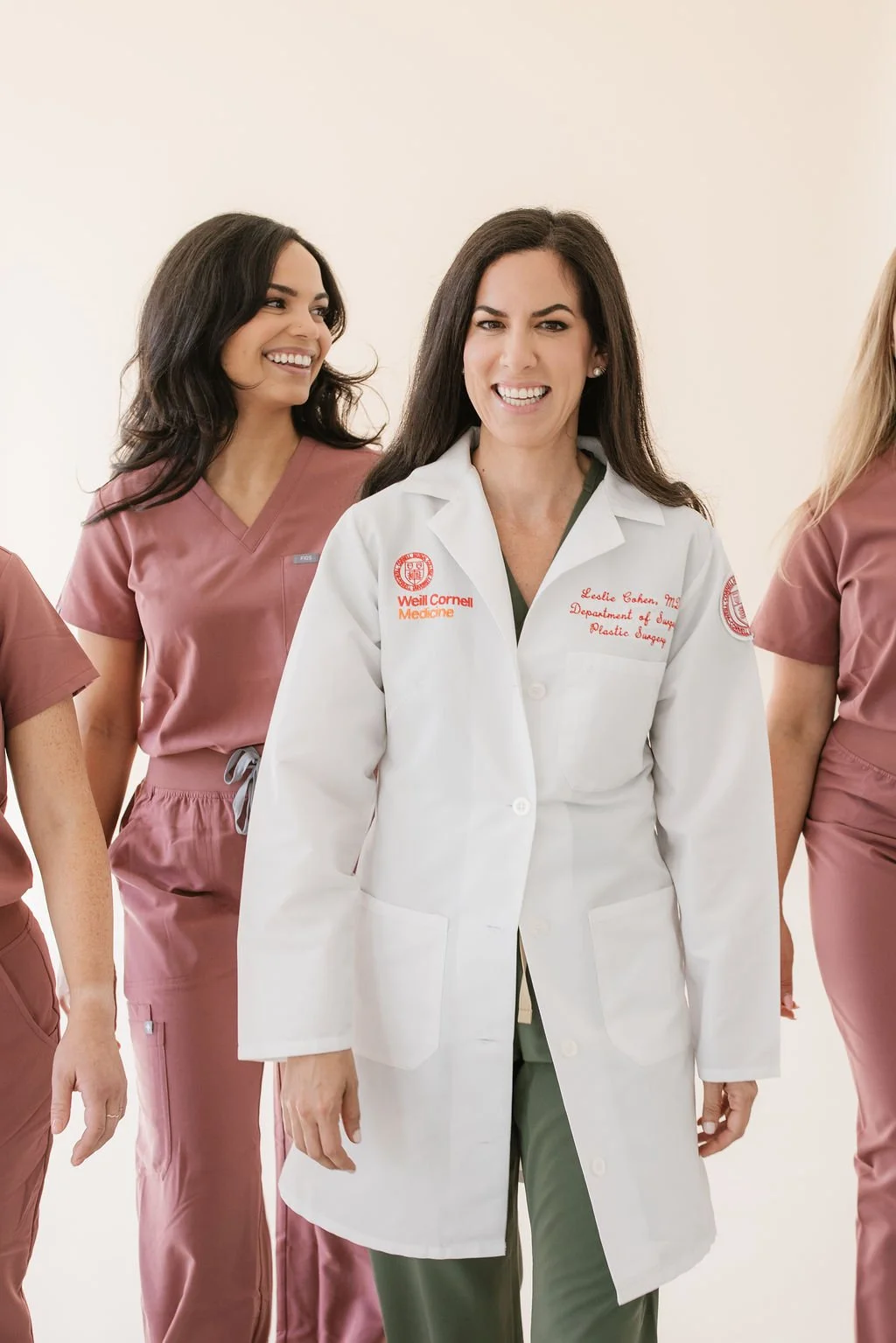 Group of female medical professionals, one in a white lab coat and others in pink scrubs, smiling and walking together.