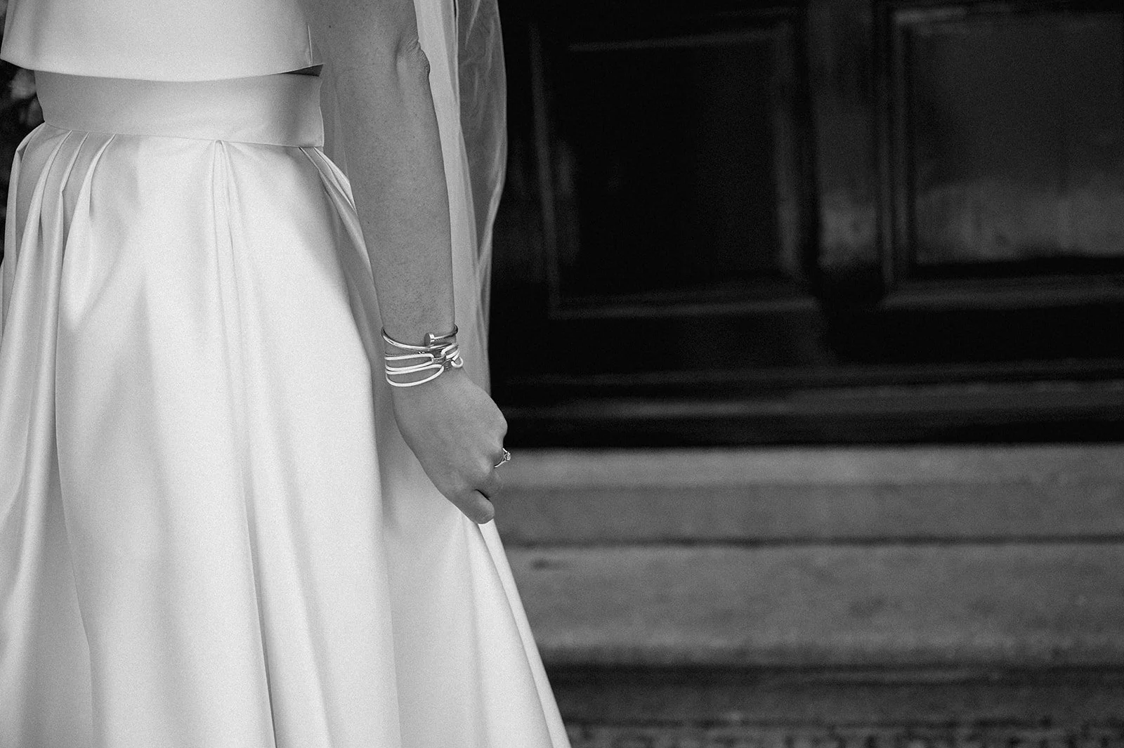 Close-up of a woman in a white wedding. dress, showing her left arm and hand with bracelets, standing near a wooden cabinet, in black and white.