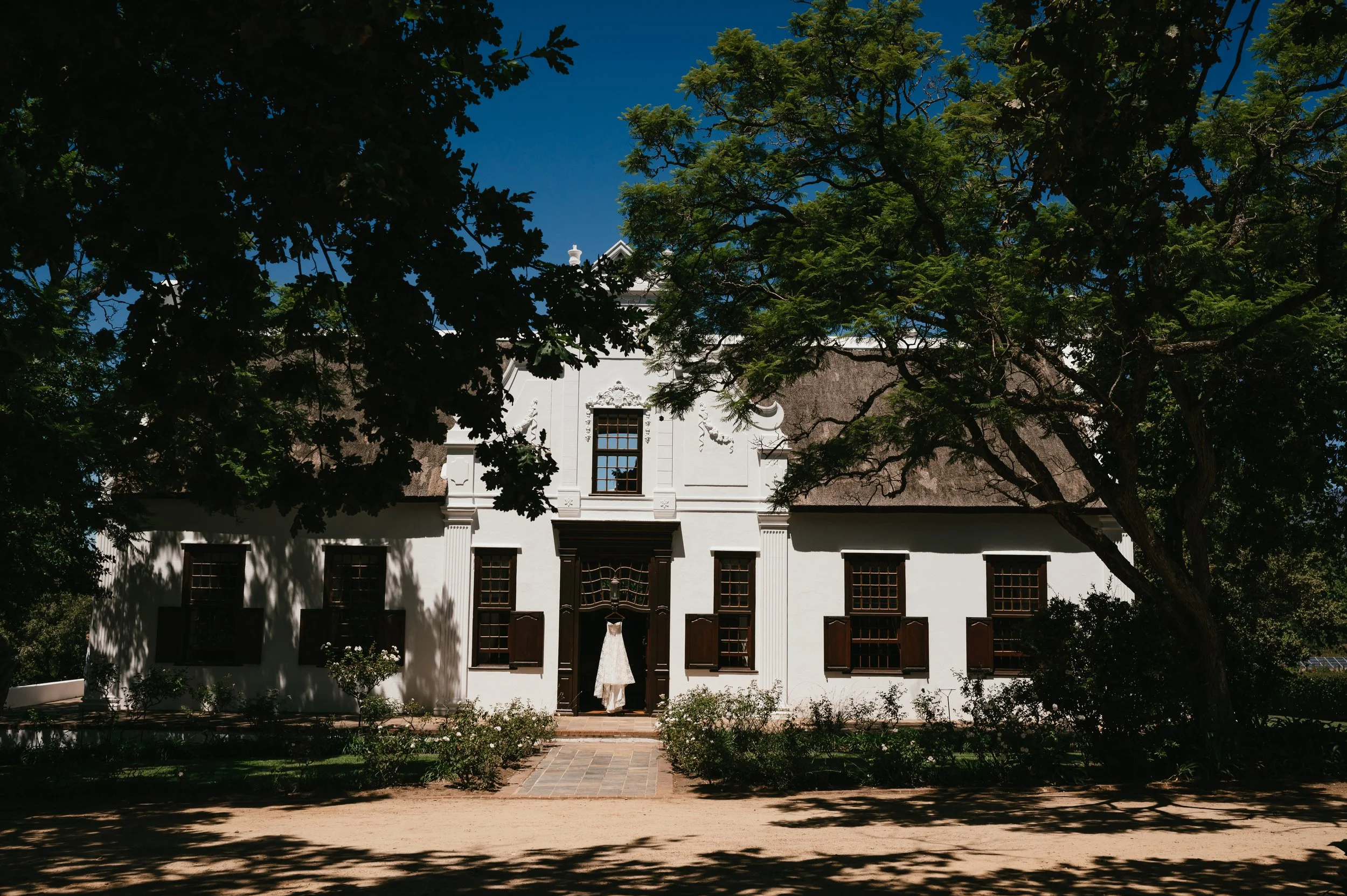 A white historic house with dark brown shutters and a thatched roof, partially obscured by large leafy trees with a clear blue sky overhead.
