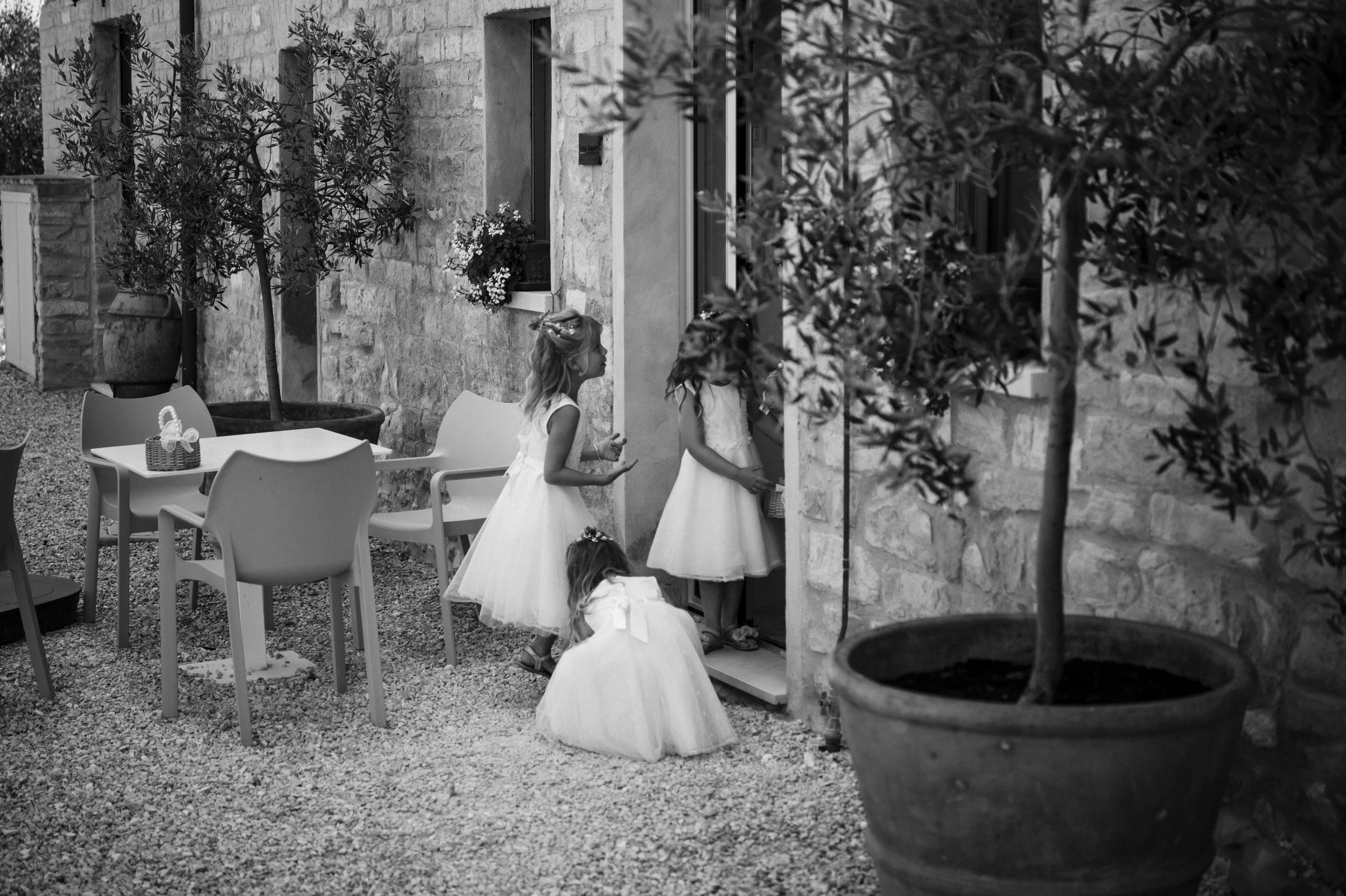Four young girls wearing white dresses standing outside a building, with one girl at the door and the others waiting, surrounded by chairs, potted plants, and a stone wall.