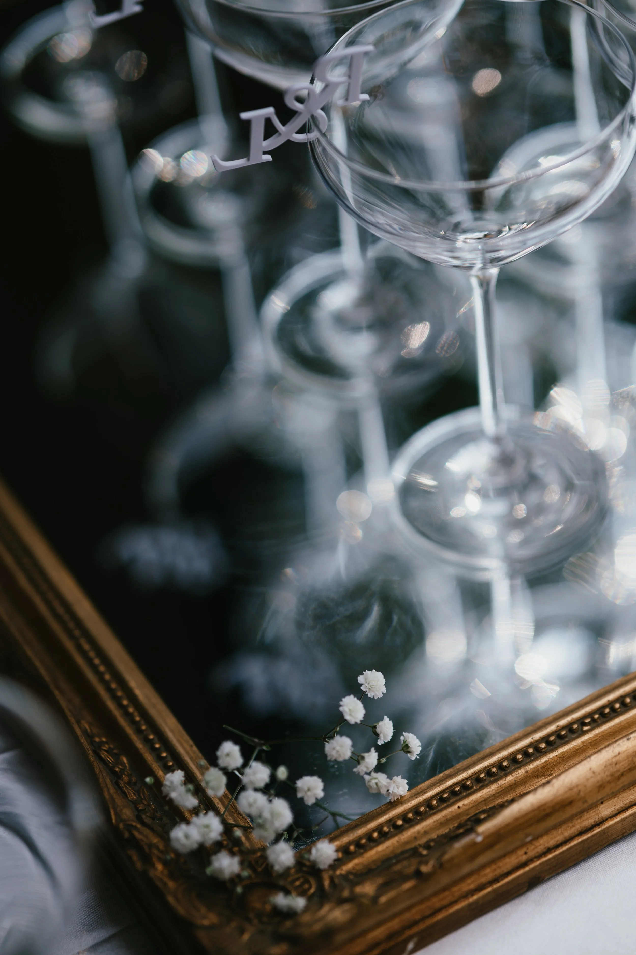 Empty wine glasses on a mirror tray with white baby's breath flowers in a gold frame, with one glass having a white letter 'A' charm hanging on it.