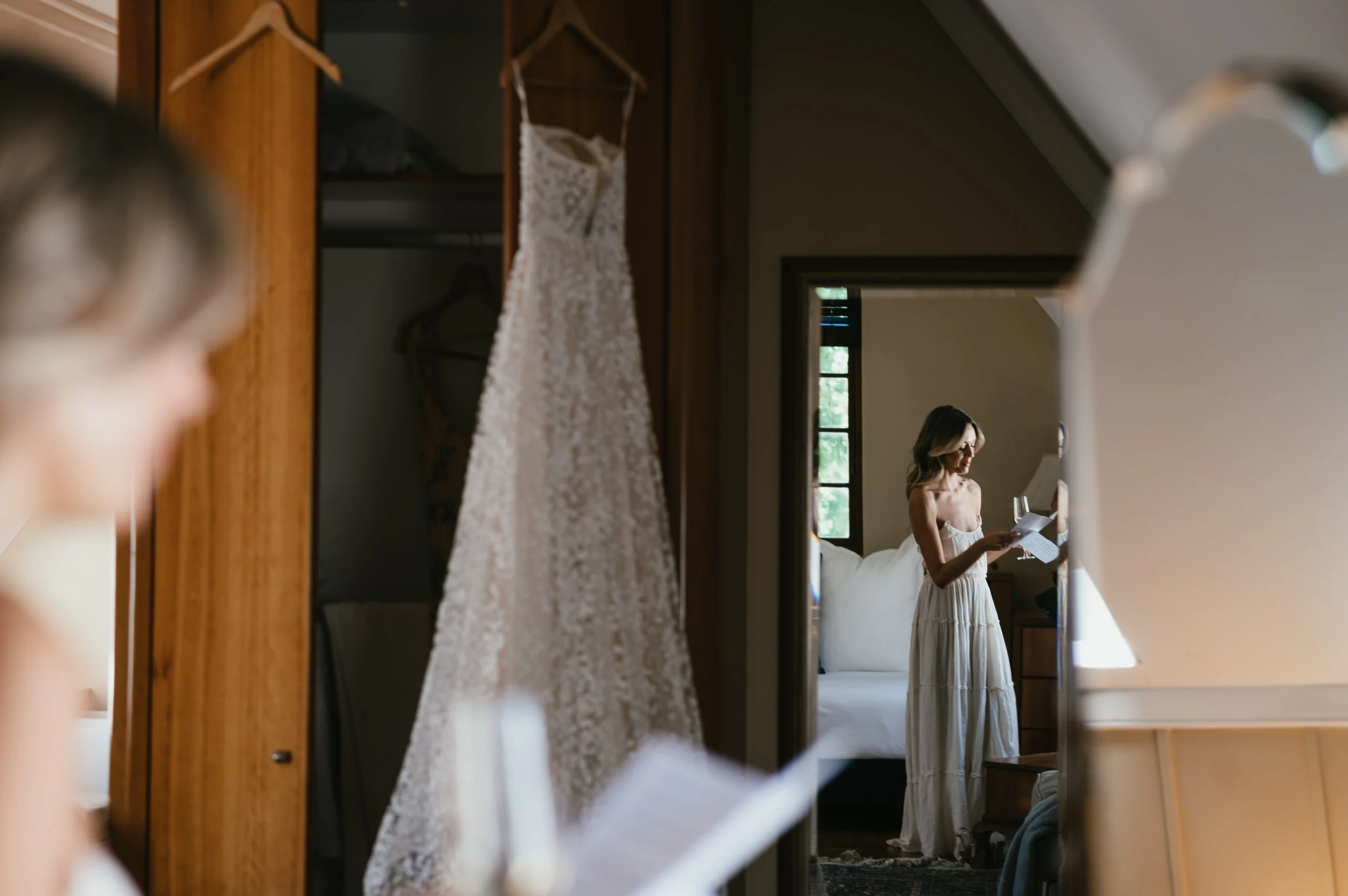 A woman in a white dress reading a letter while standing by a bed, seen through a mirror in a bedroom with wooden furniture and a wedding dress hanging.
