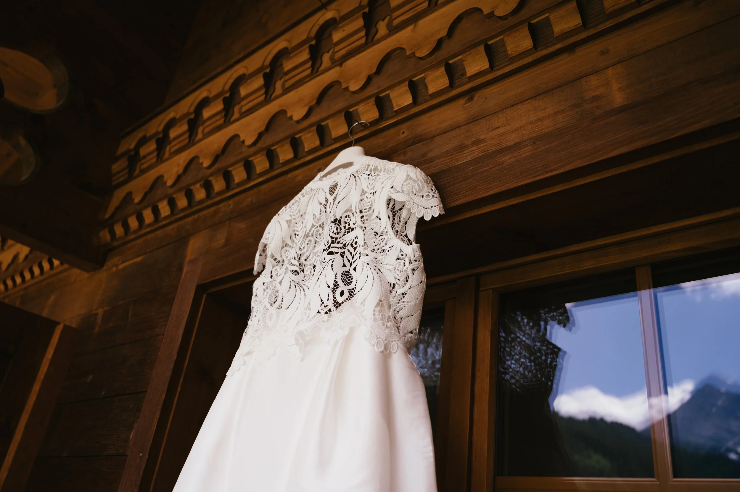 A white wedding dress with intricate lace detailing hanging on a hanger inside a wooden room with a window reflecting a mountain and trees.