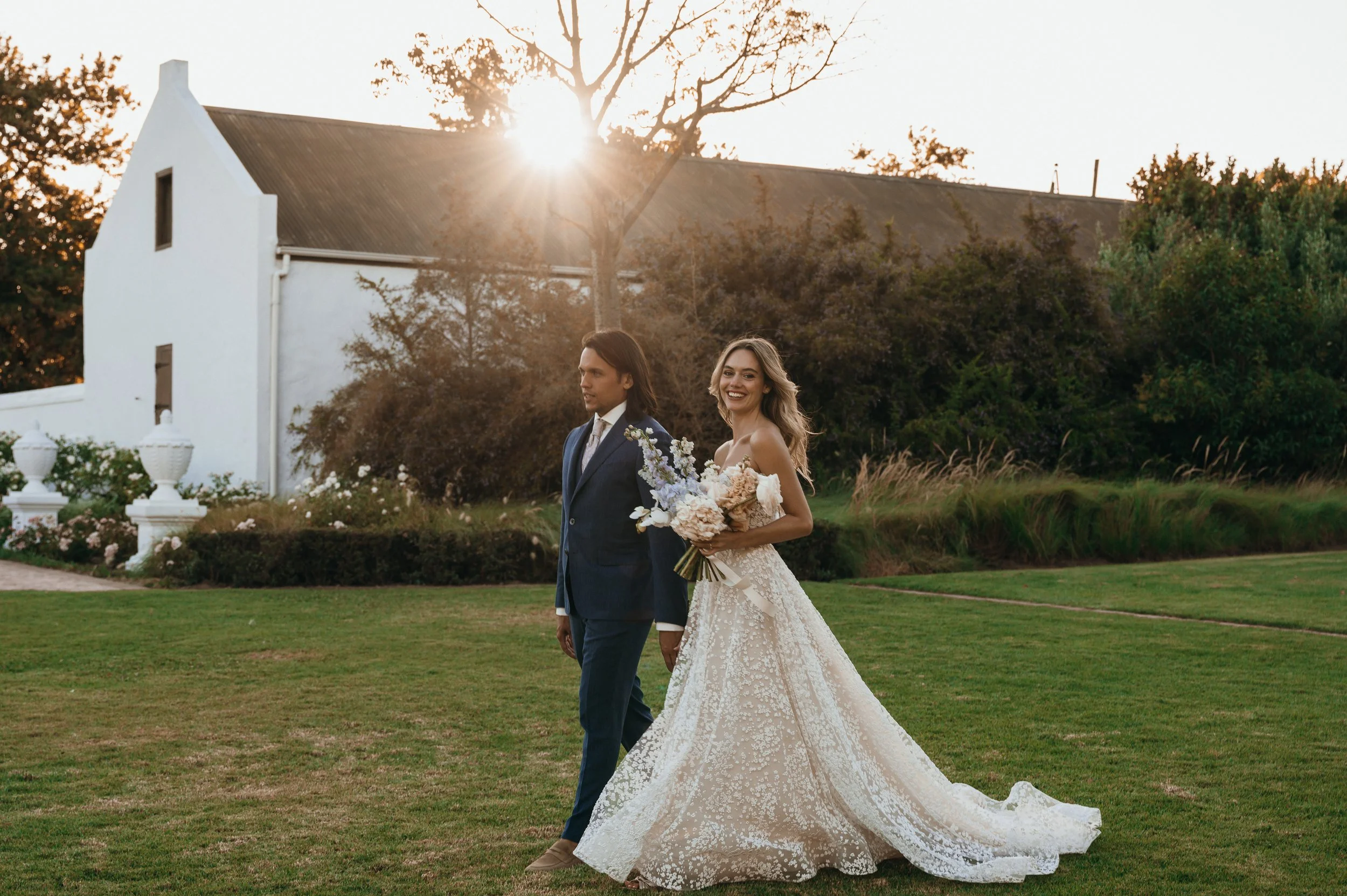 A bride and groom walking hand in hand outdoors during sunset. The bride is smiling and holding a bouquet, wearing a lace wedding gown. The groom is in a navy suit and tie. There is a white building and trees in the background.