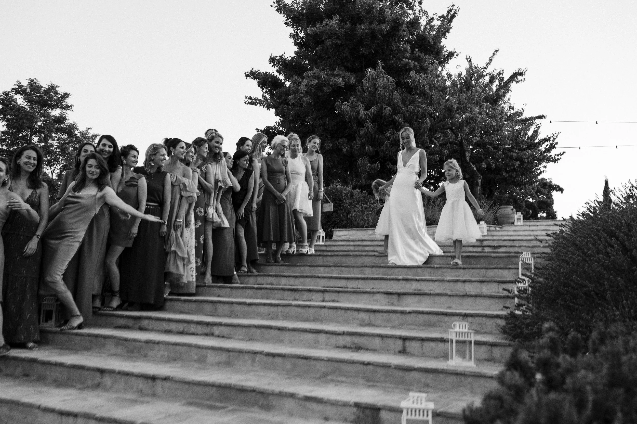 A bride walking down steps with a young girl, surrounded by a group of women, at an outdoor wedding ceremony in the evening.