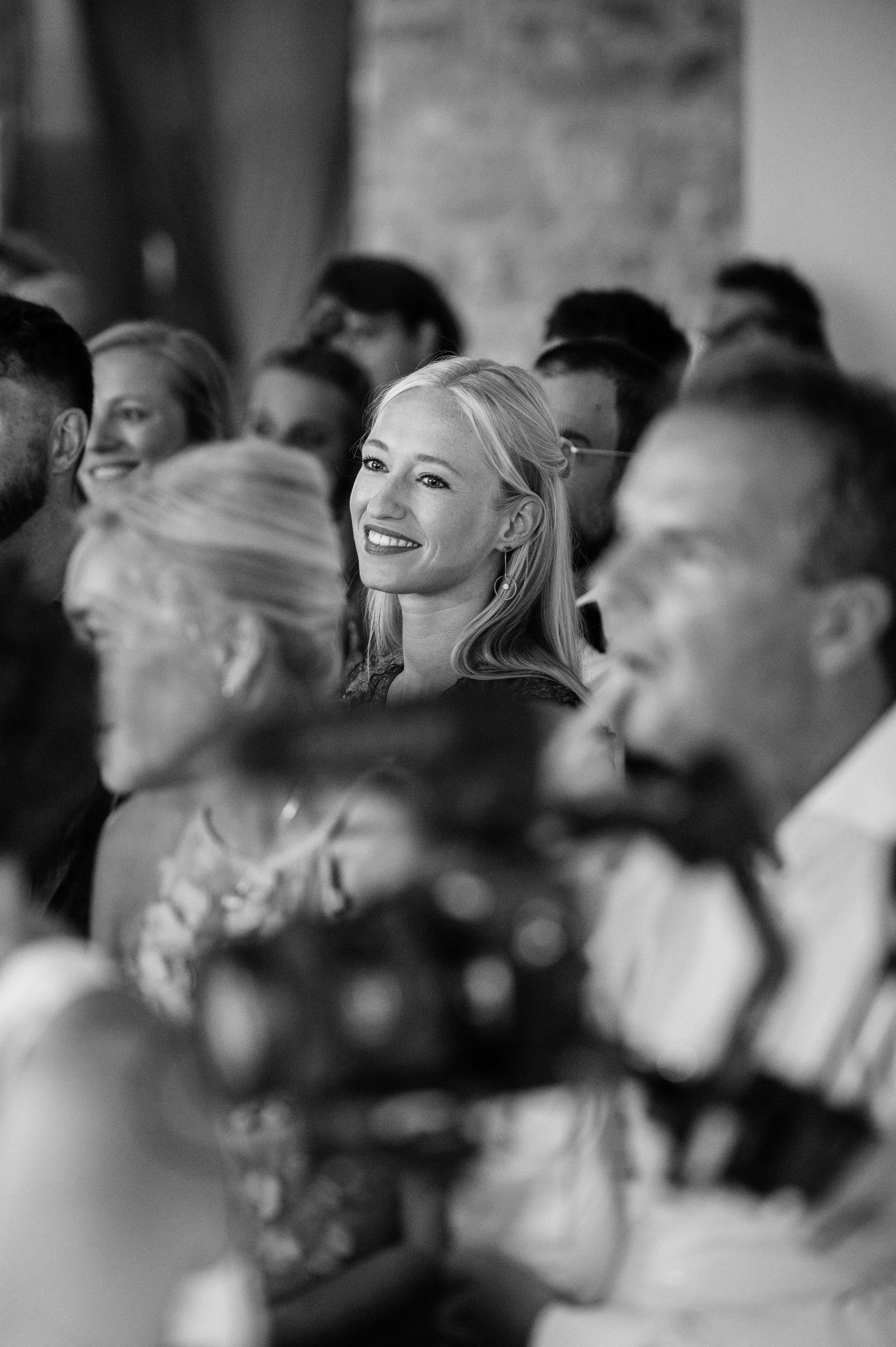 A group of people seated at an event, some smiling and looking ahead, with a woman with blonde hair and earrings in the center smiling.