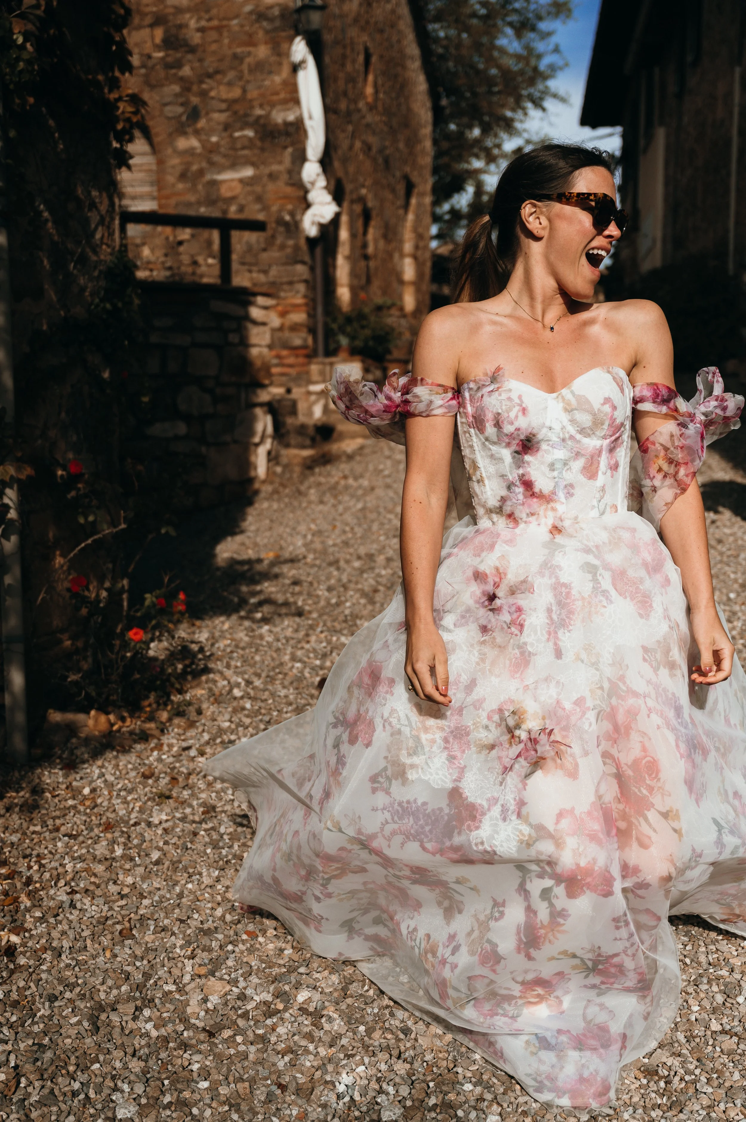 A woman in a floral wedding dress laughing outdoors on a gravel path.