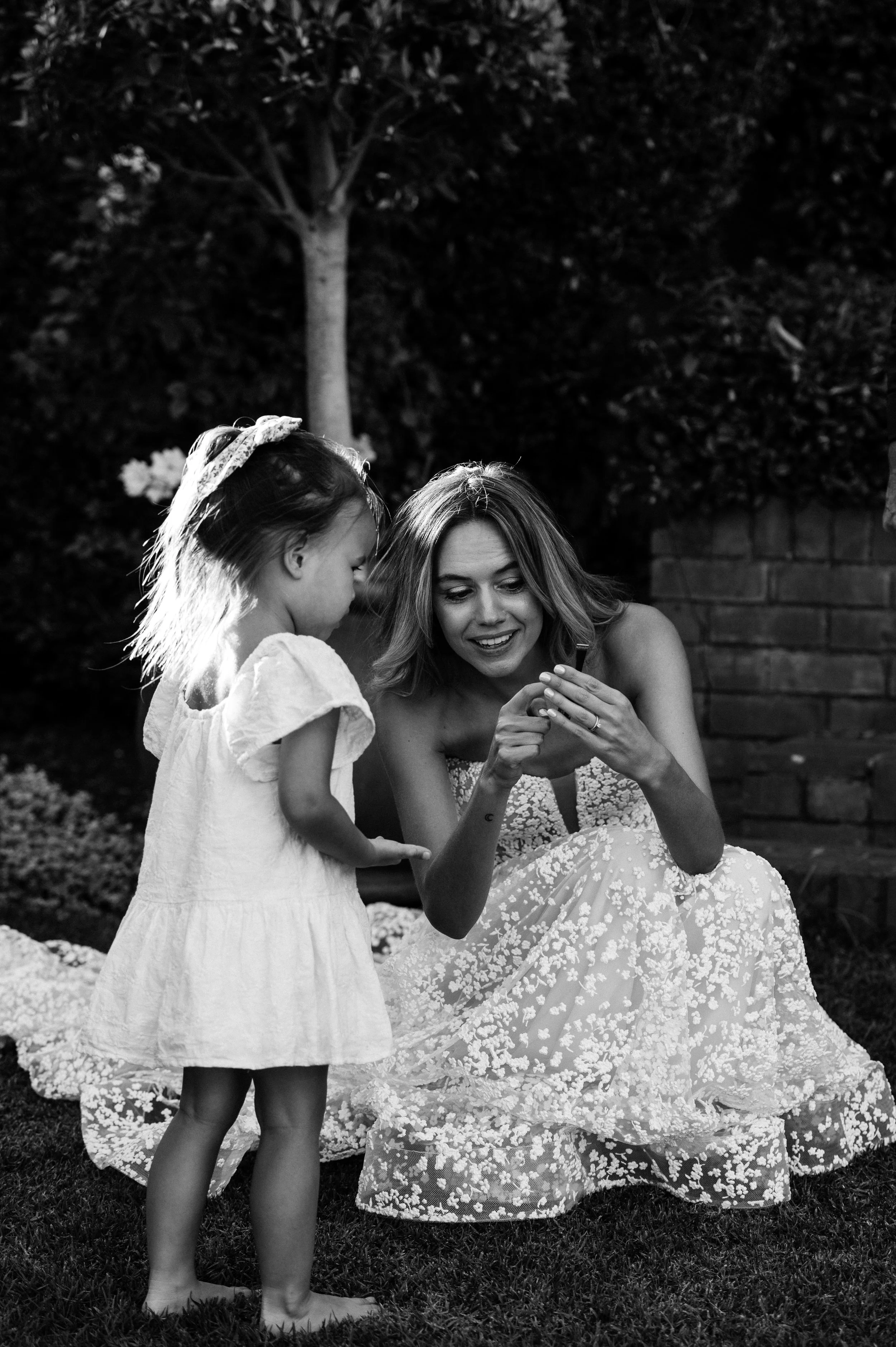 A woman in a wedding dress showing something on her phone to a young girl in a white dress outdoors at night.