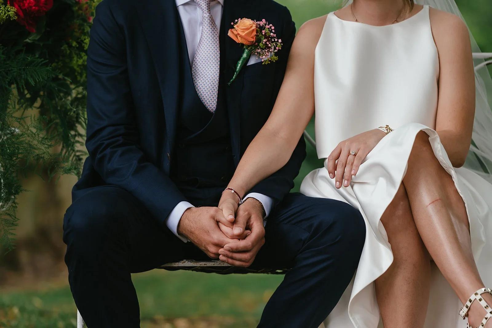 Close-up of a bride and groom sitting on a swing, holding hands. The groom is wearing a dark suit with a boutonniere, and the bride is in a white dress with a ring on her finger, outdoors with greenery in the background.
