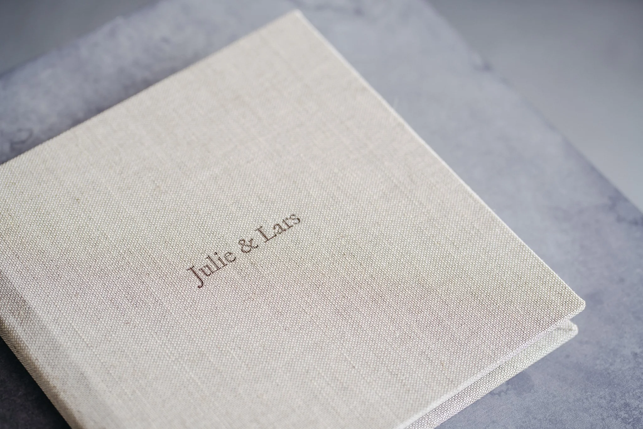 A close-up of a cream-colored fabric-covered box with the words 'Julic & Lais' embroidered in brown on the lid, resting on a gray surface.