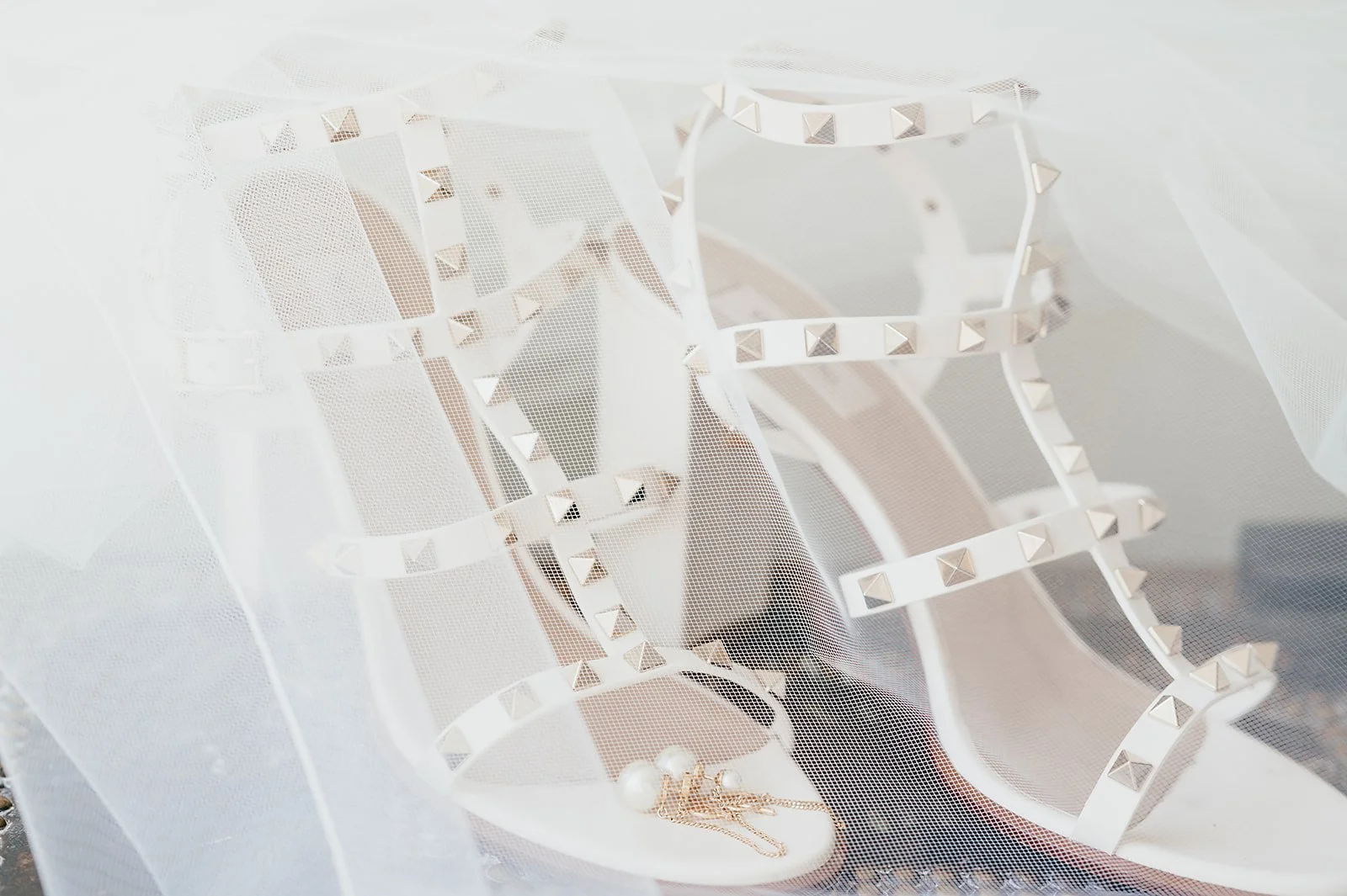 White high-heeled shoes with studded straps, a pearl necklace, and earrings behind a sheer white fabric with small holes.