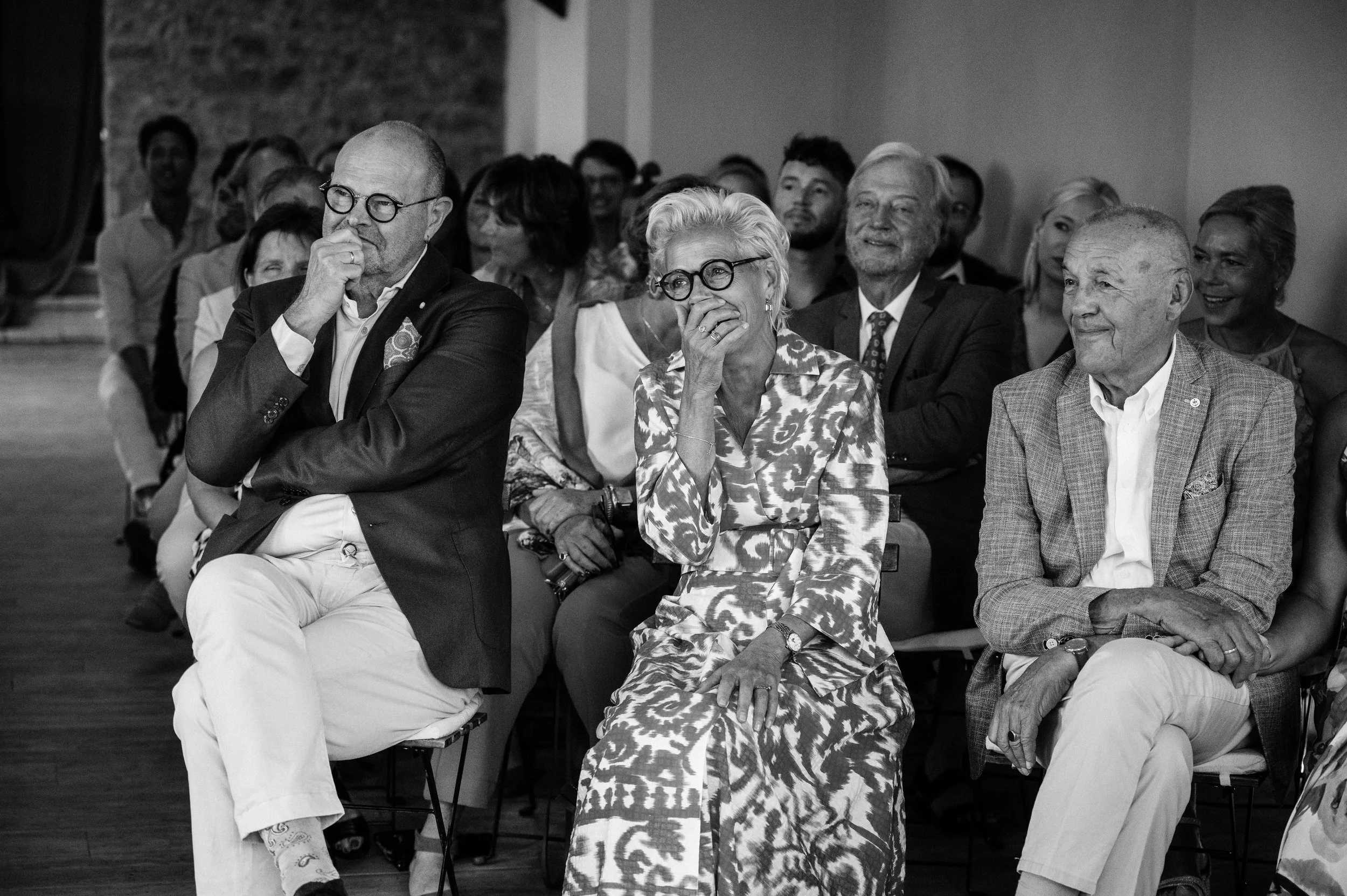 Black and white photo of an audience at a formal event, showing three prominent individuals in front row: a bald man with glasses wearing a dark blazer and light pants, a woman with short white hair wearing glasses and a patterned dress, and an older