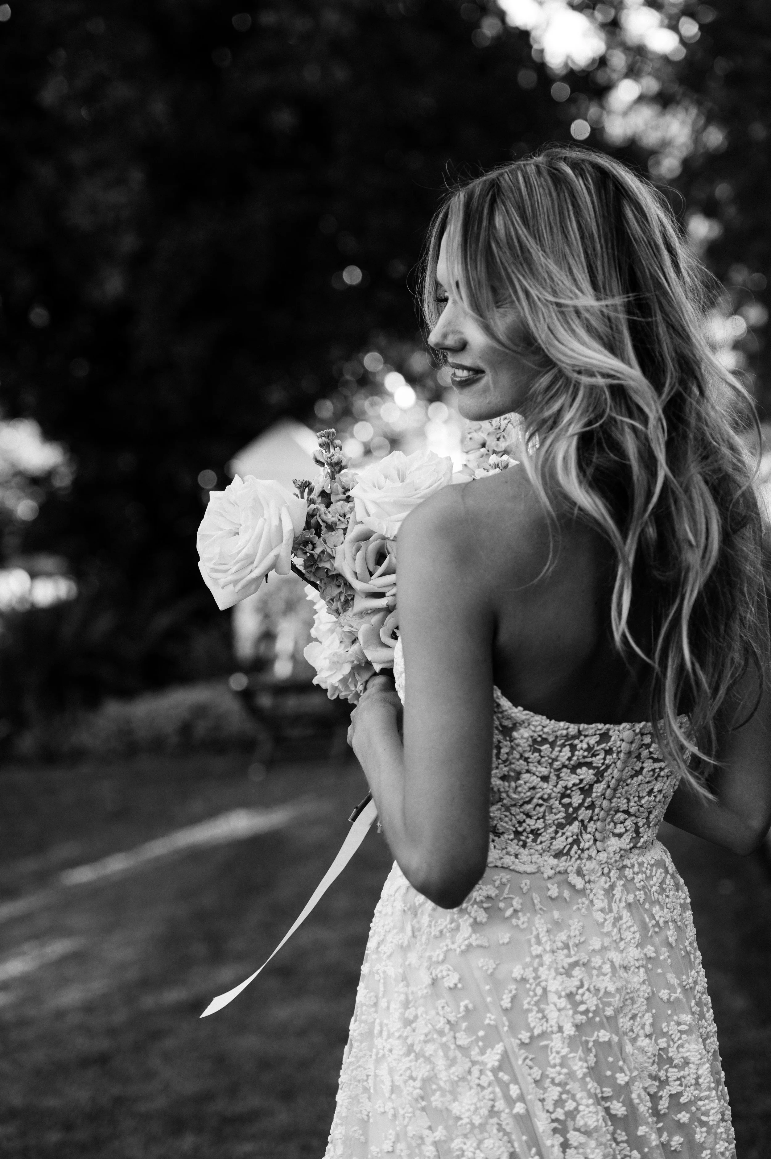 A woman in a wedding dress holding a bouquet of flowers outdoors, smiling with her eyes closed.