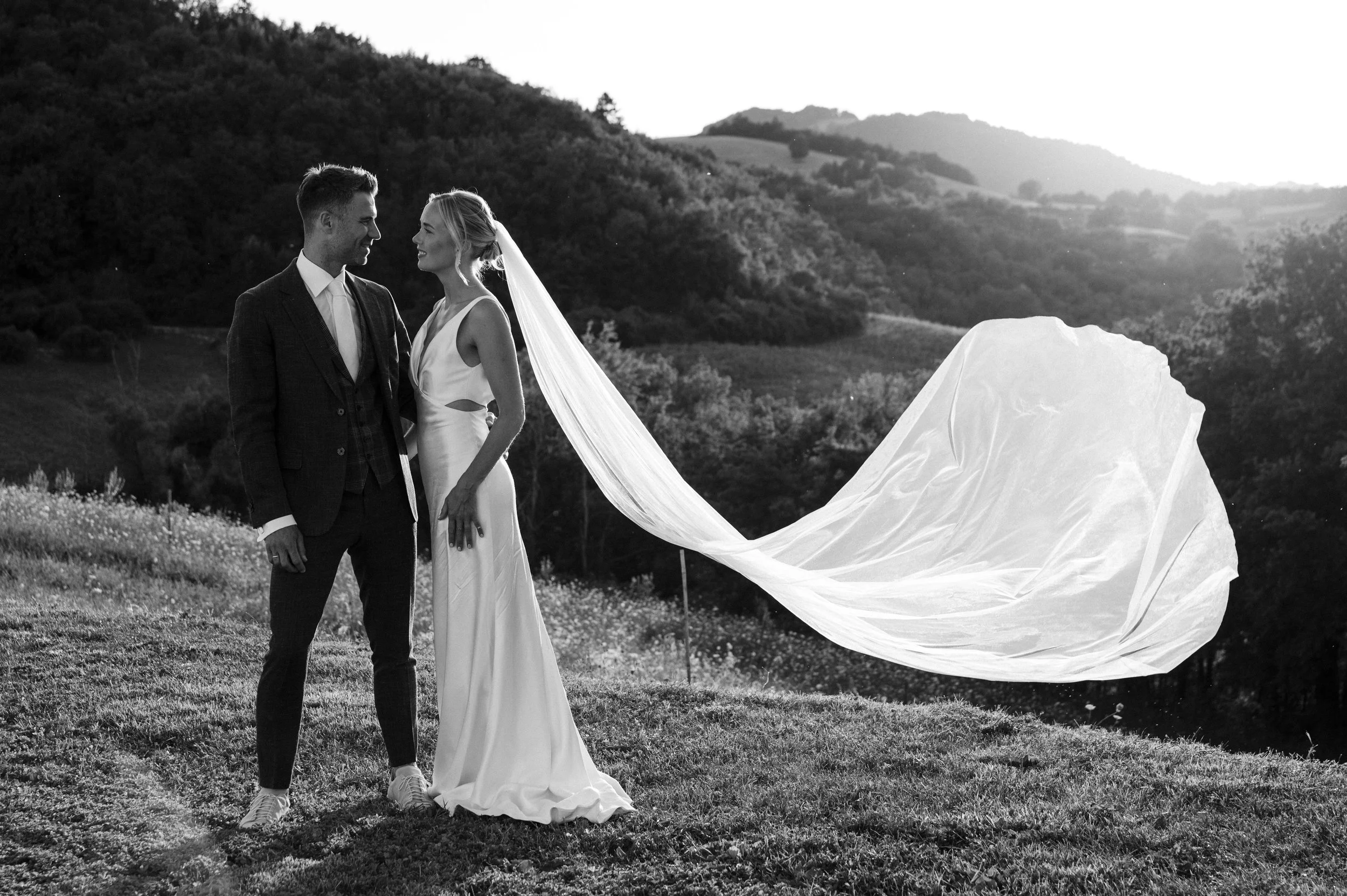 A bride and groom standing in a grassy field with rolling hills in the background, sharing a moment on their wedding day, with the bride's long veil flowing behind her.