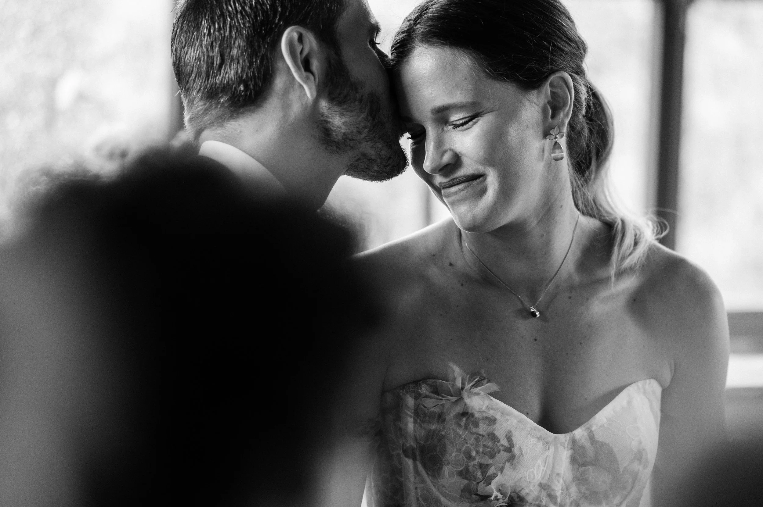 A black and white photo of a woman with a bride's dress and a necklace, and a man whispering in her ear, both smiling with closed eyes.