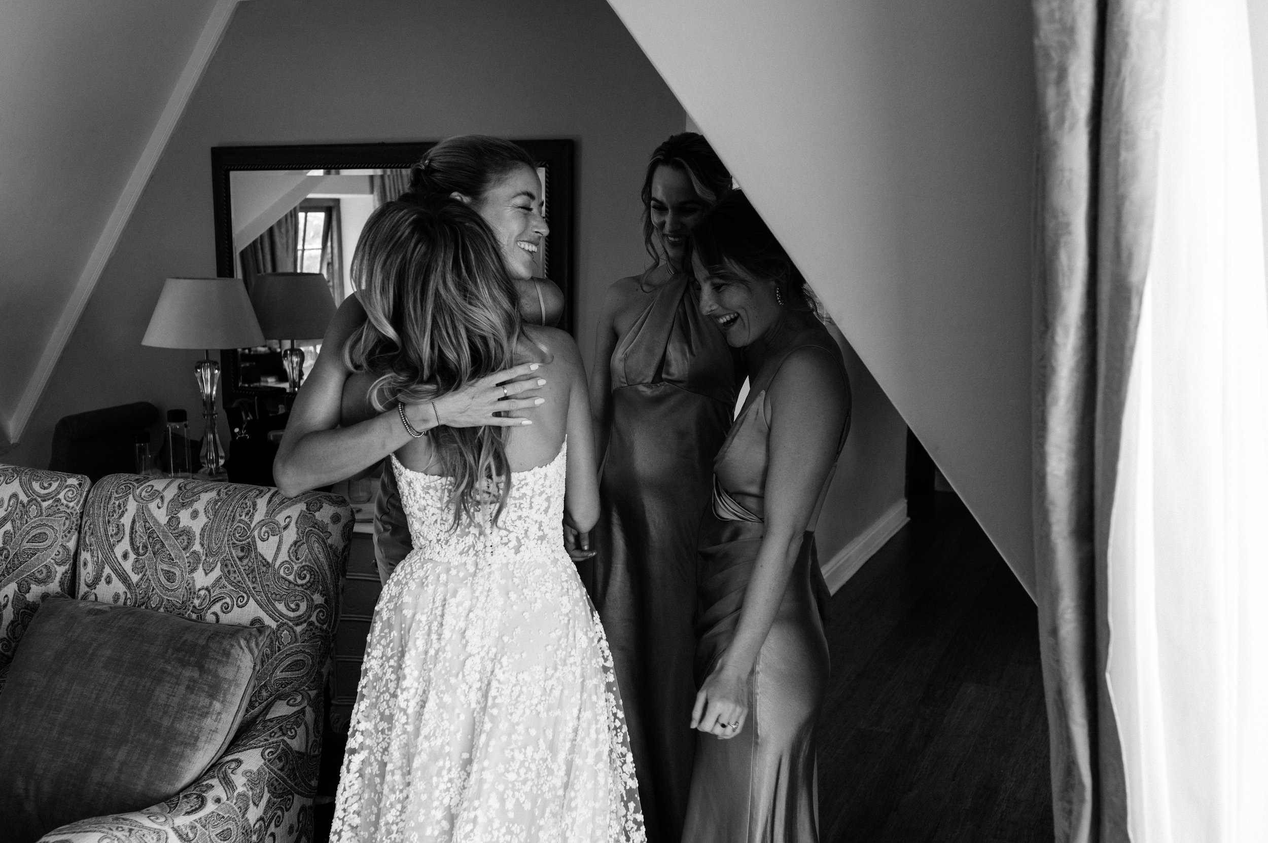 Four women in elegant dresses sharing a joyful moment and hugging each other in a cozy room.