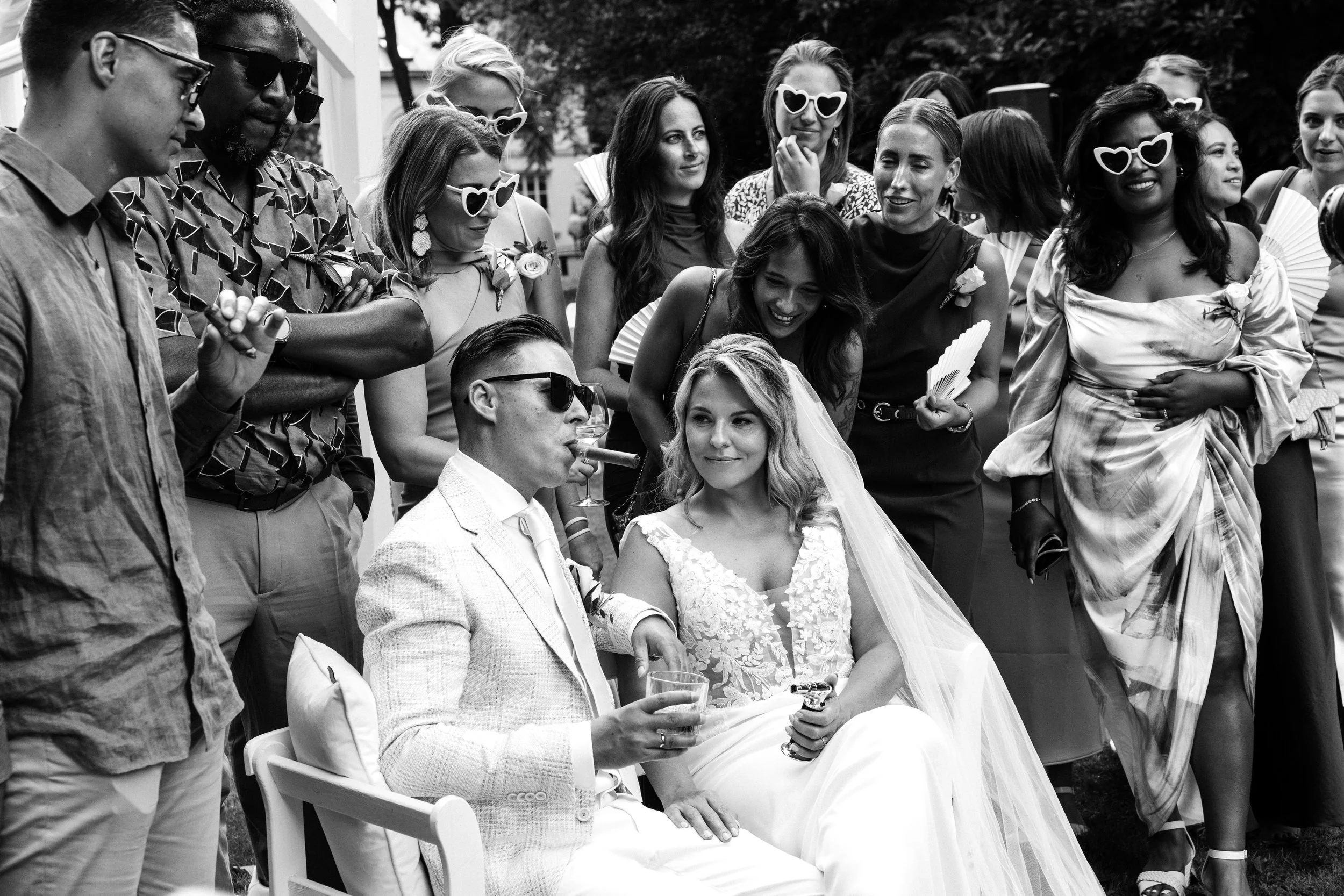 A black and white photo of a wedding celebration with a bride and groom sitting on a bench, surrounded by guests. The groom is wearing sunglasses and is smoking a cigar while holding a drink, and the bride is smiling, wearing a wedding dress and veil