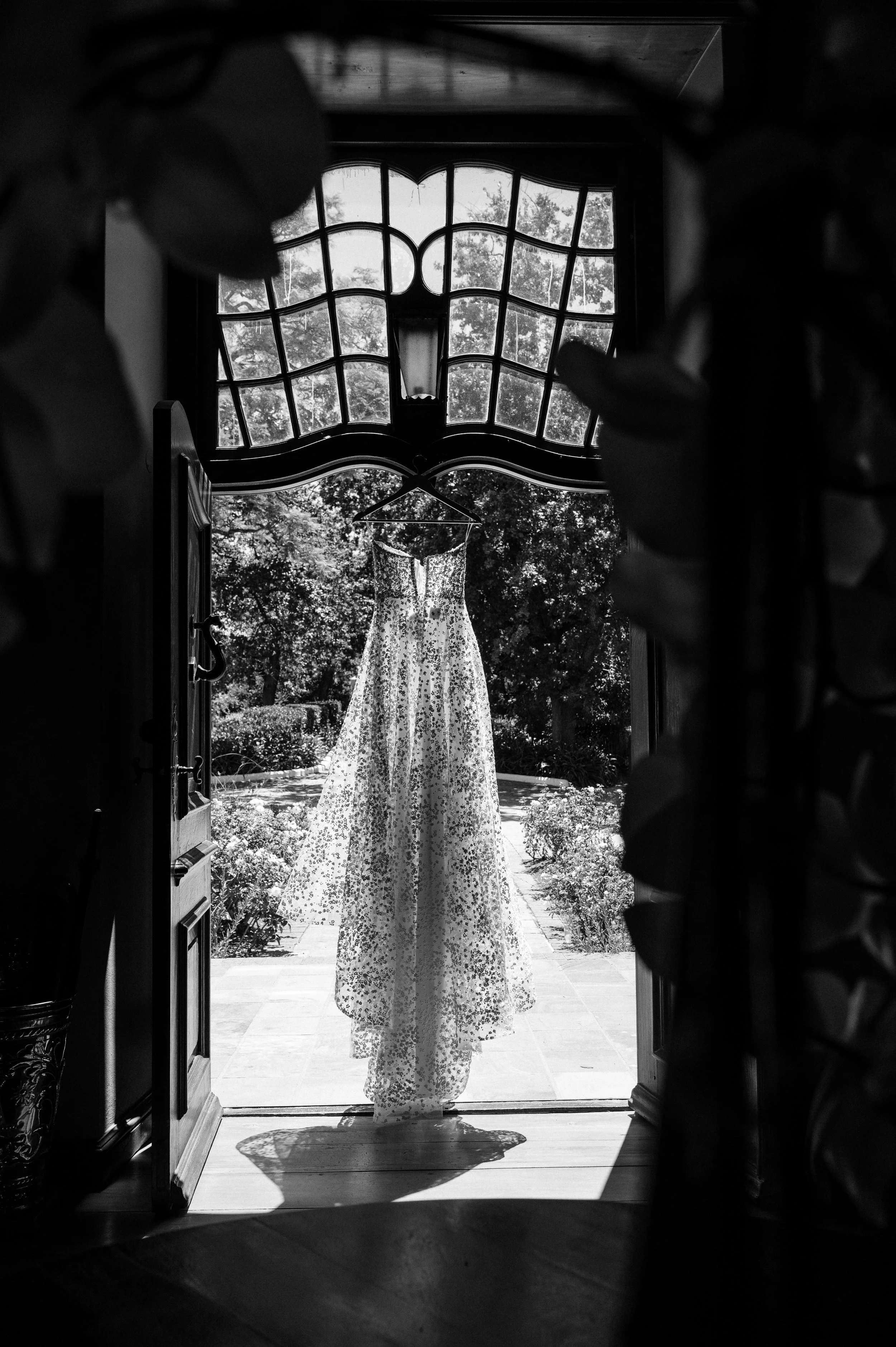 A wedding dress hanging outside a doorway with trees and plants in the background, viewed through an ornate window.