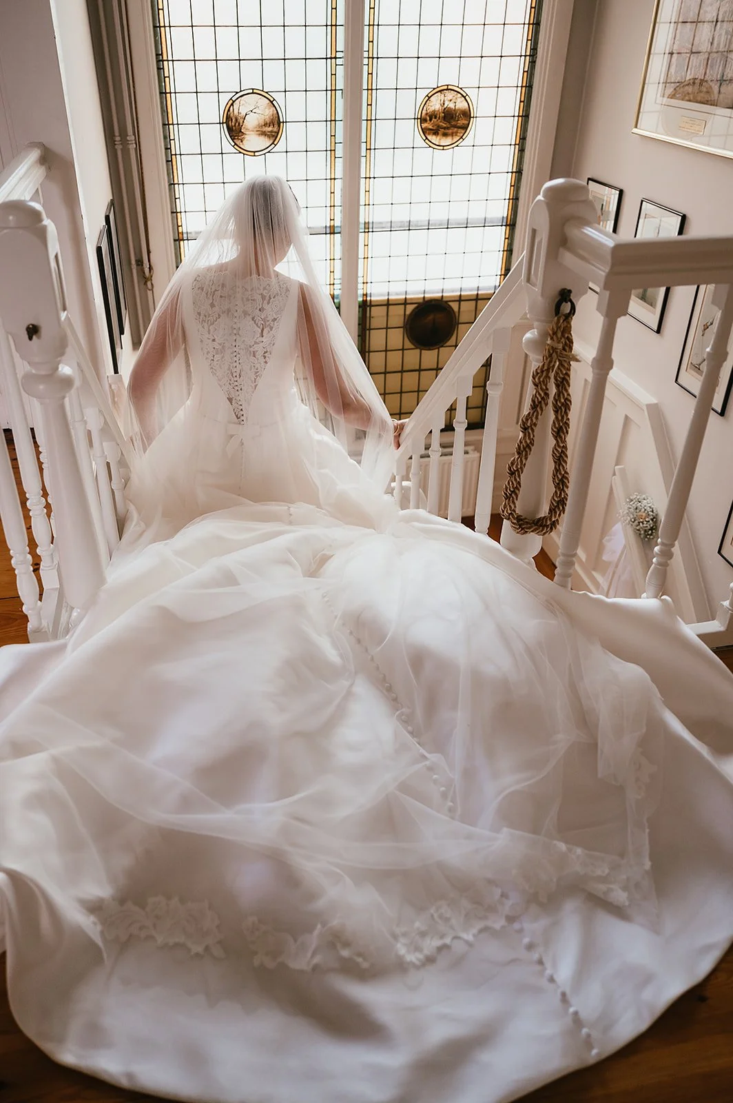 A bride in a white wedding gown with a lace back and a long train, walking down a staircase with a veil covering her head, in a well-lit indoor setting.