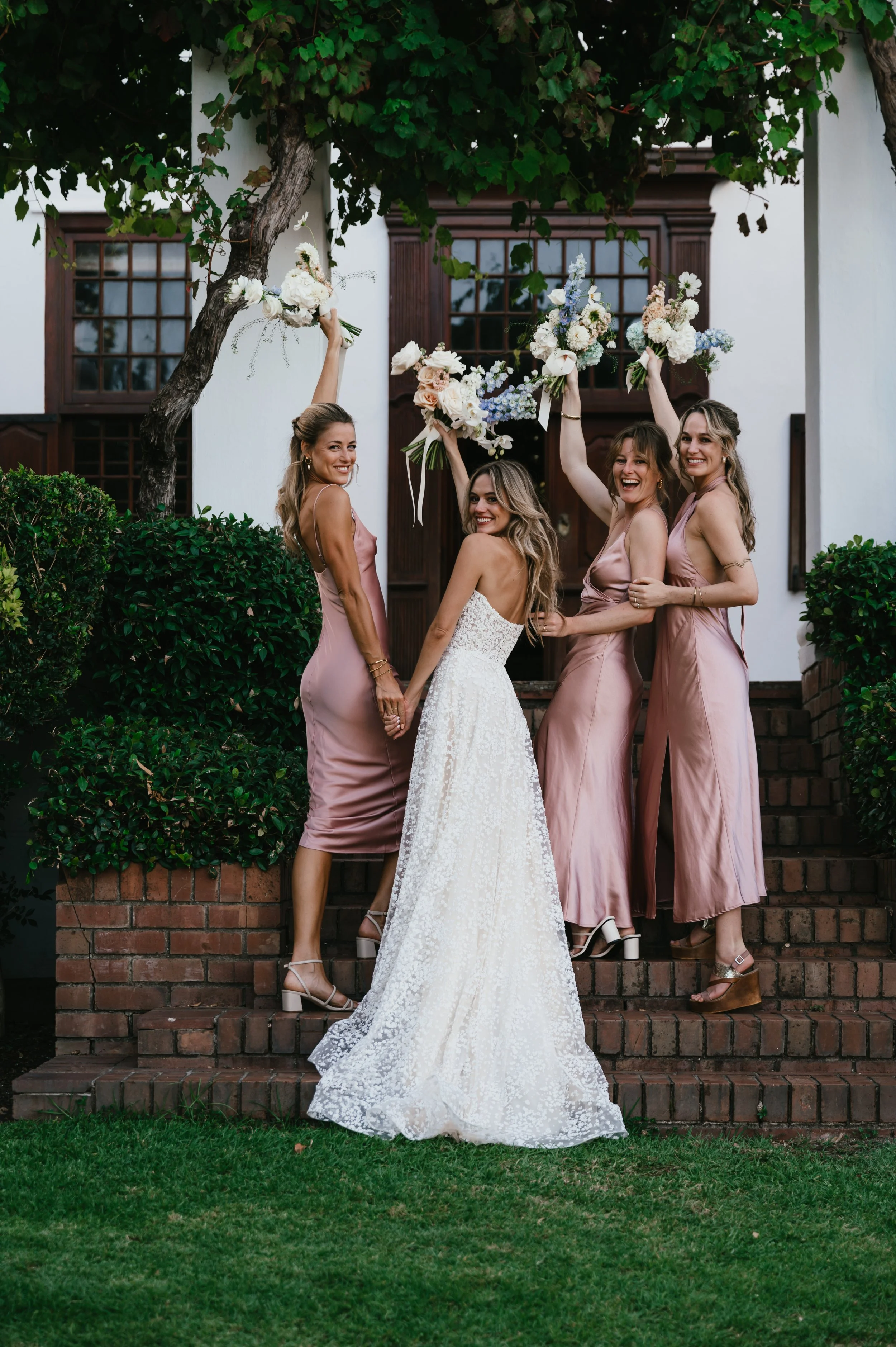 Four women, one bride in a white wedding gown and three bridesmaids in pink dresses, happily posing on a brick staircase outdoors with green bushes and a white house in the background. The bridesmaids are holding bouquets in the air.