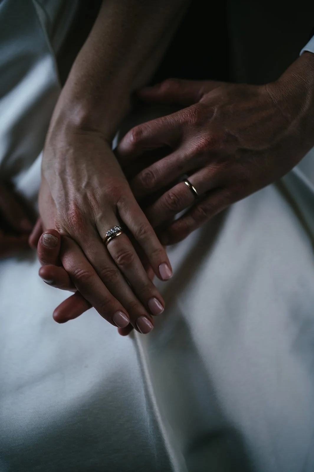 Close-up of two people’s hands, likely at a wedding or engagement, with rings on their ring fingers, resting on a surface.