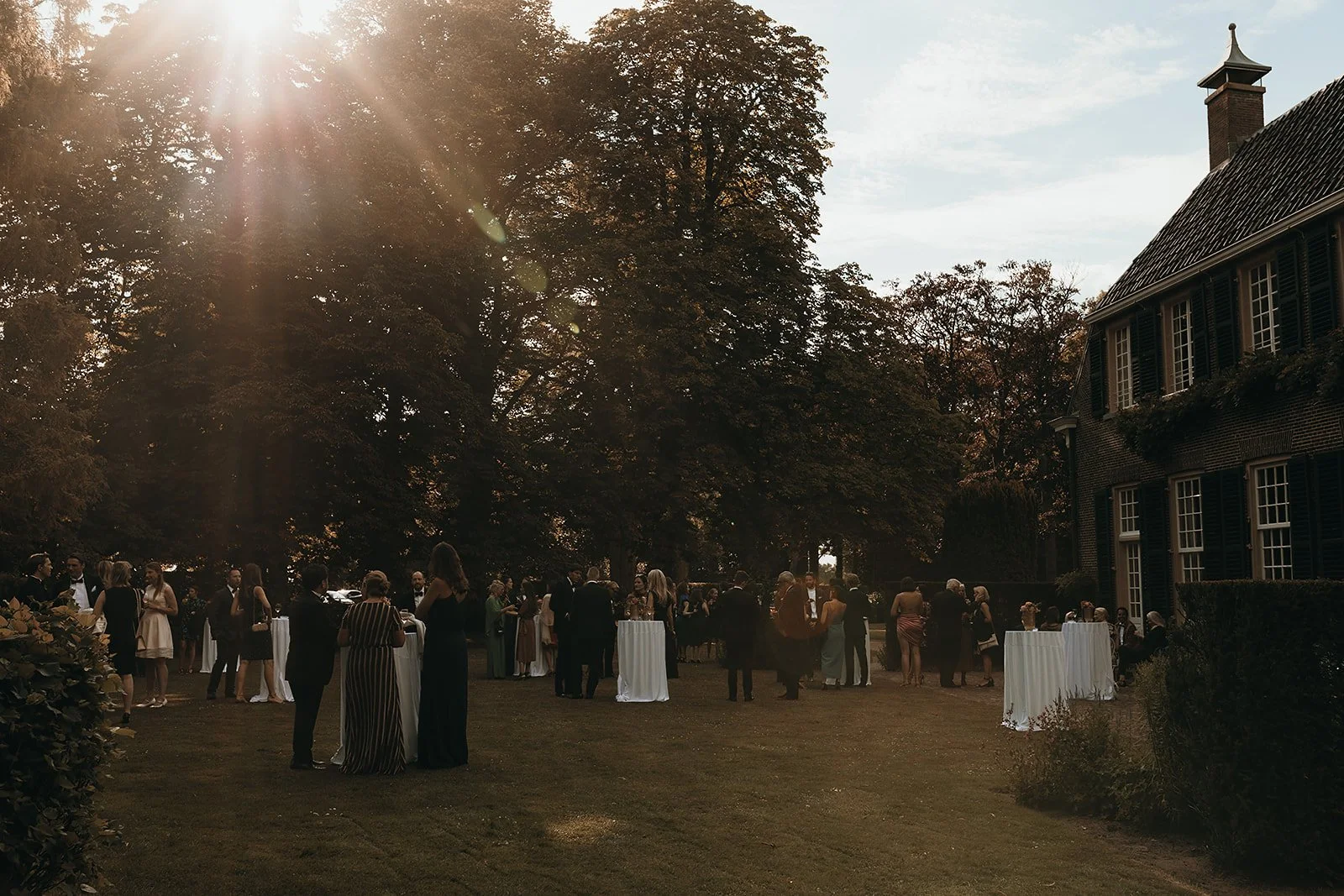 A gathering of people in formal attire at an outdoor event on a lawn with a large tree and a brick house with green shutters in the background, under a sunny sky.