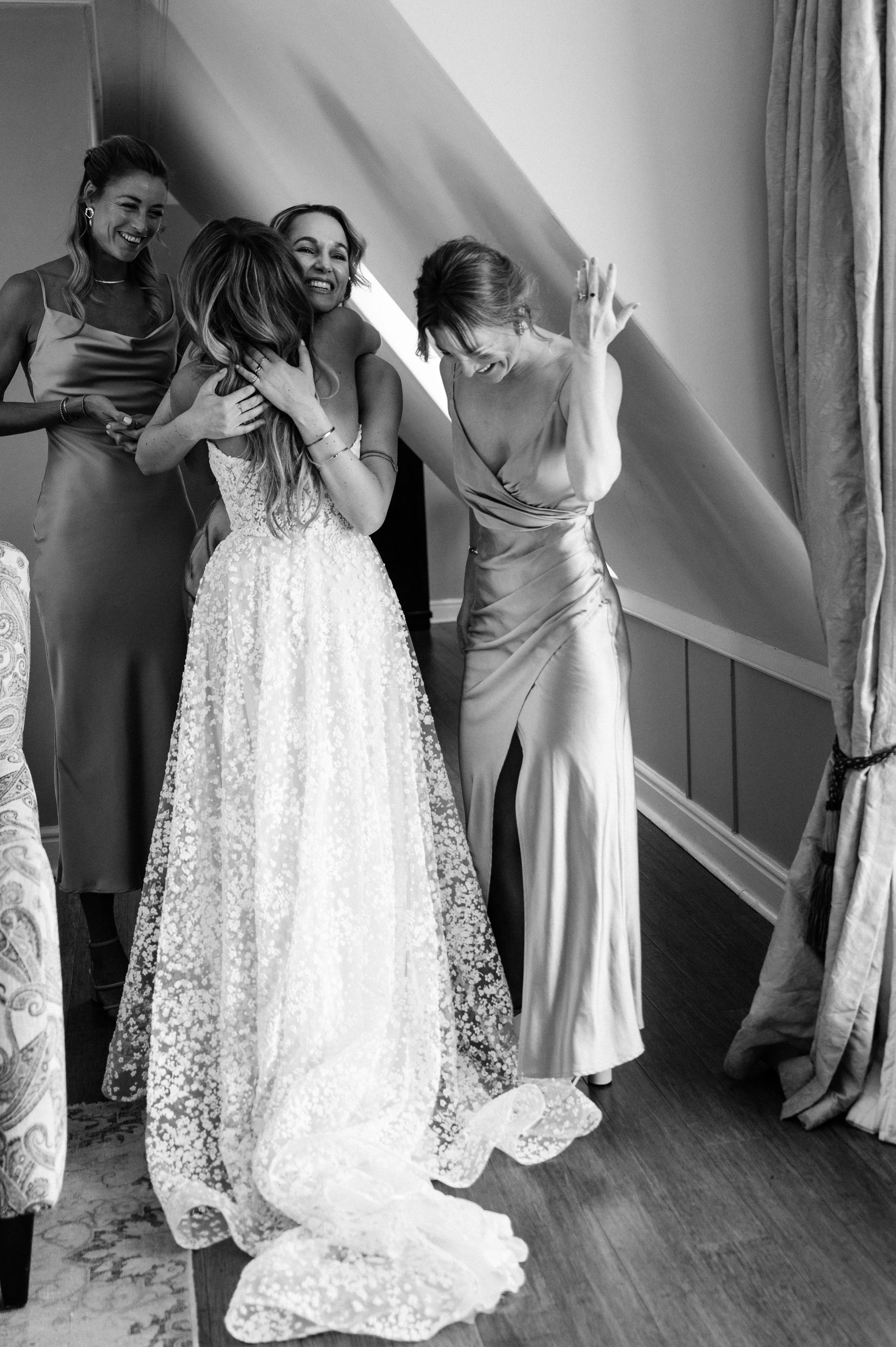 A joyful group of women, including a bride in a wedding dress, sharing a hug and laughs in a room with a sloped ceiling and curtains.