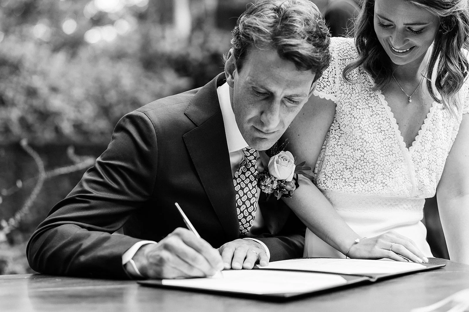 A black and white photo of a groom signing a document with a woman standing beside him, both smiling, during a wedding ceremony outdoors.