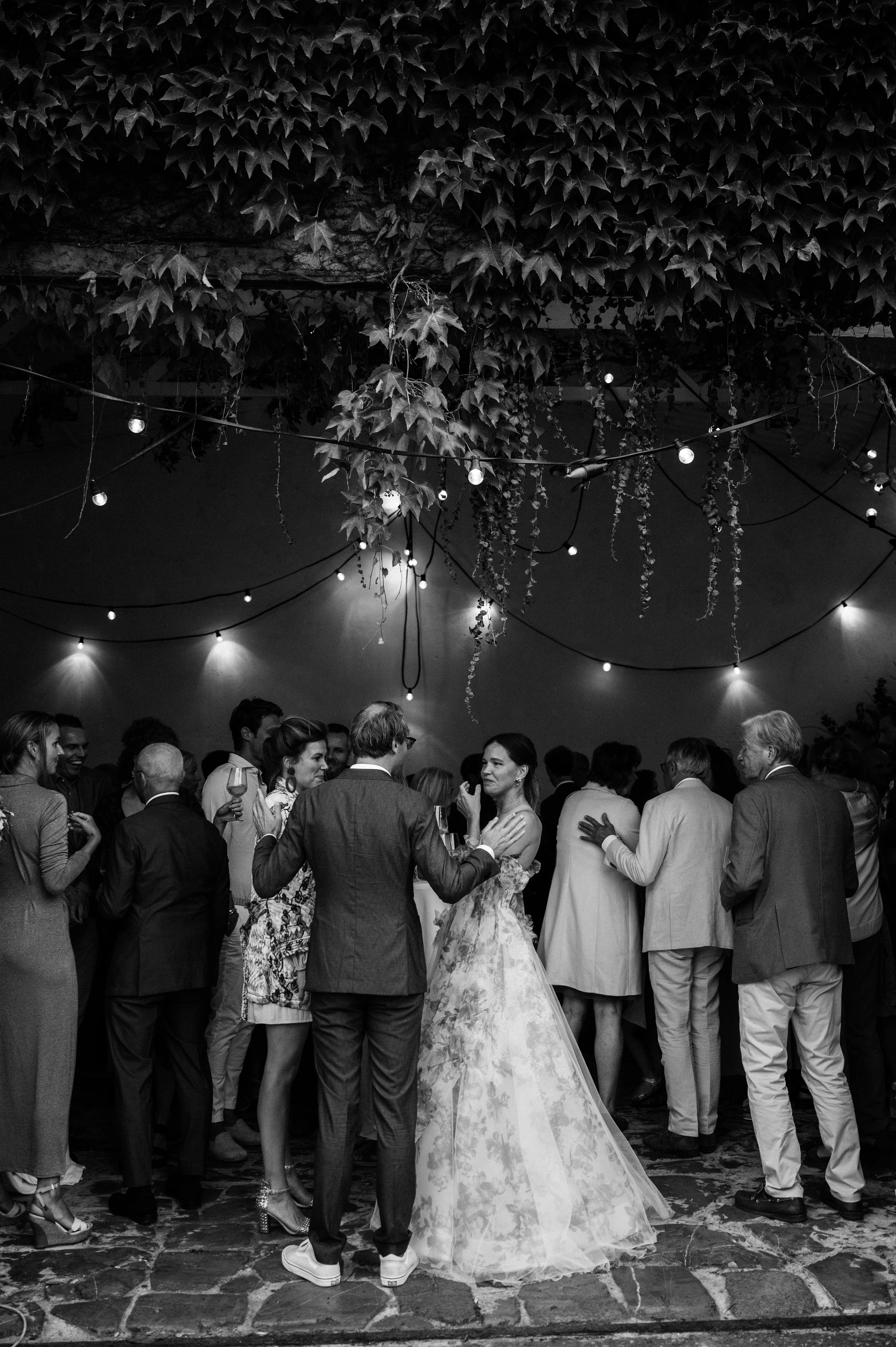 A black and white photo of a wedding reception with a bride in a gown and groom dancing with guests, under hanging string lights and leafy decorations.