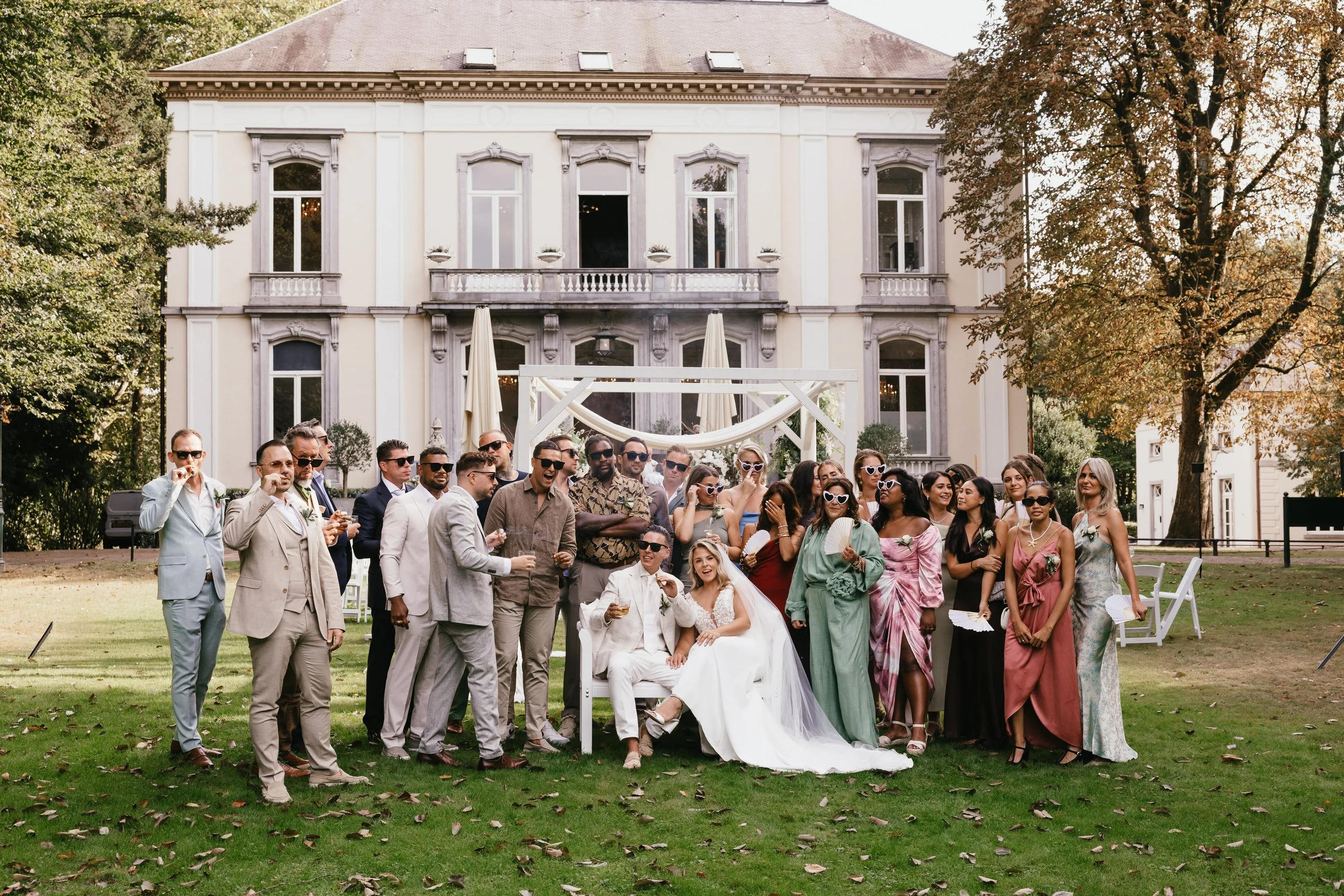 Group of wedding guests and a bride and groom in front of a large white building, outdoors on grass with trees, during daytime.