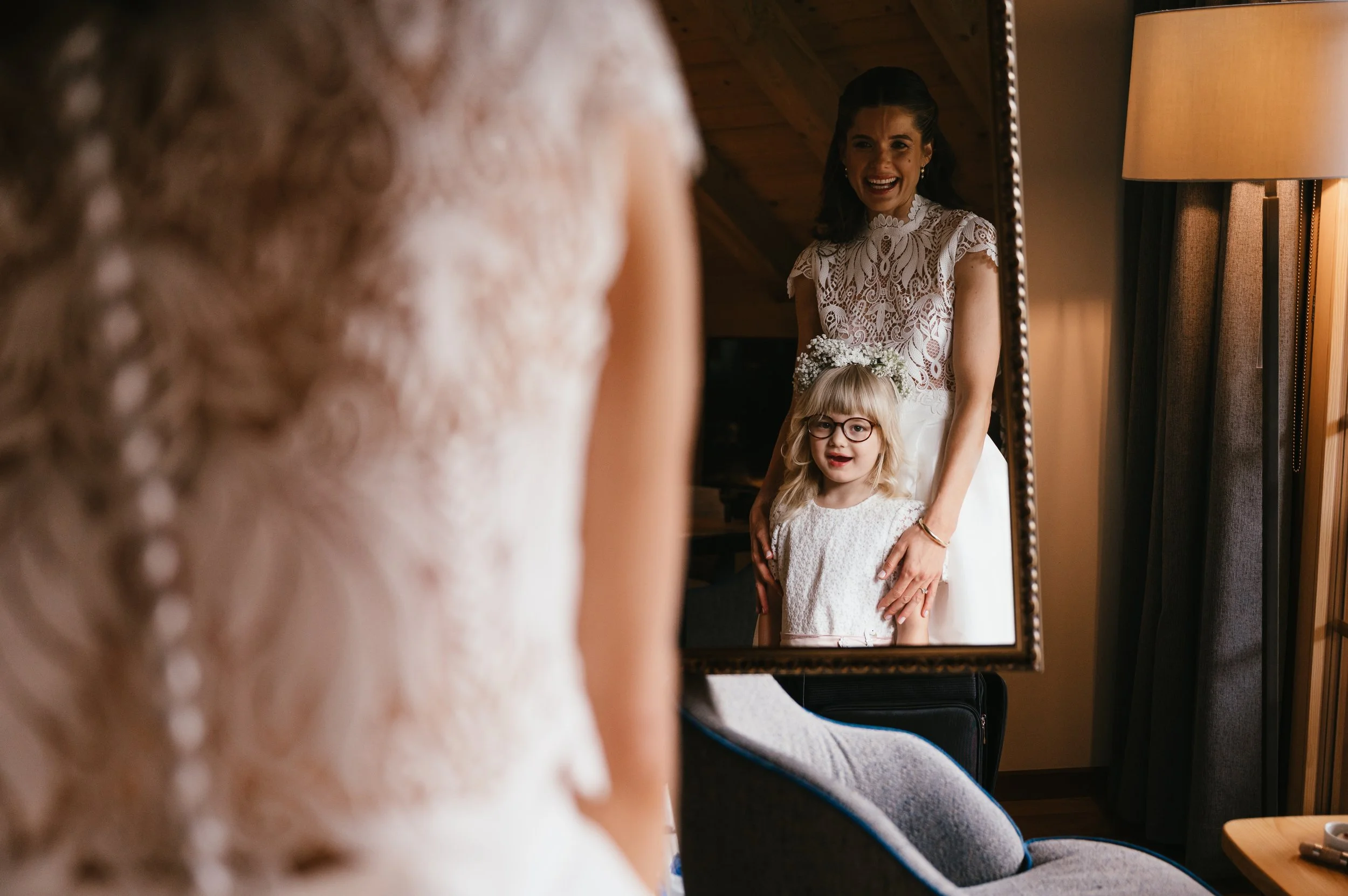 A woman in a lace wedding dress looks at her reflection in a mirror, smiling with her hand on a young girl with blonde hair and glasses, wearing a white dress and floral headband.
