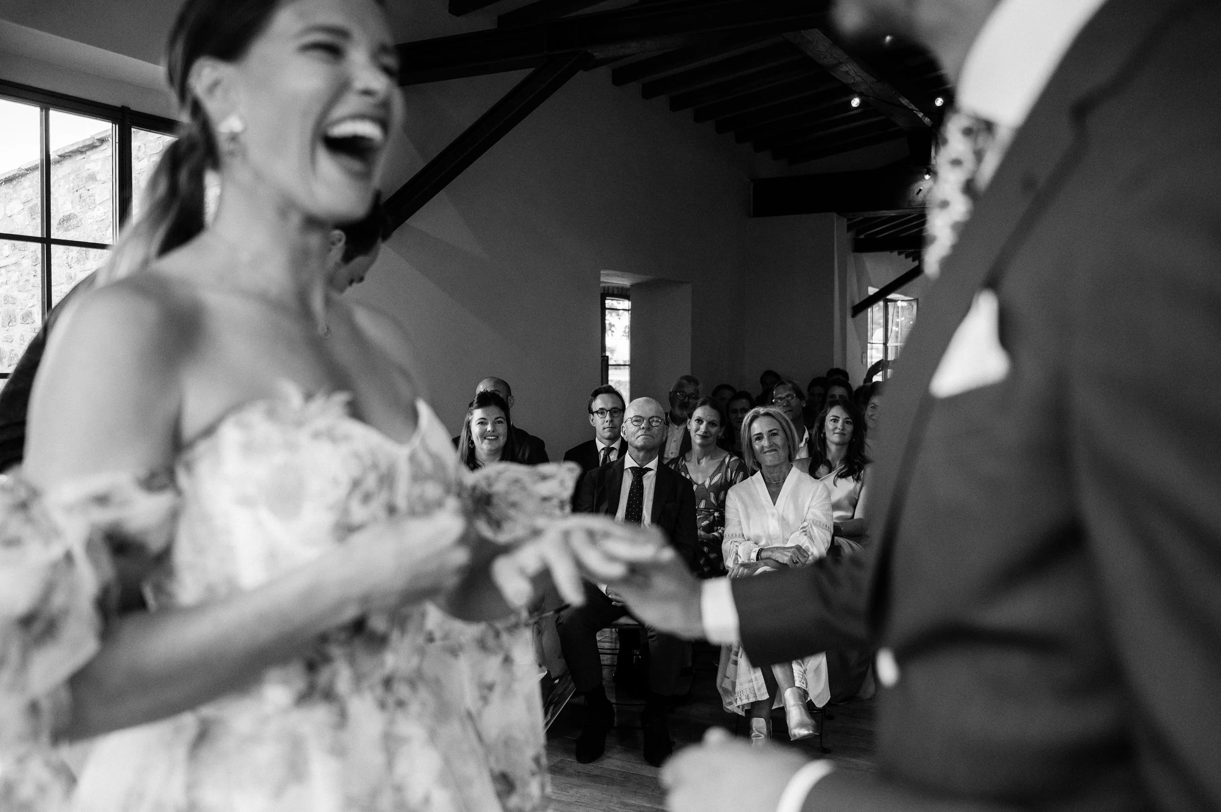 A black and white photo showing a woman in a wedding dress smiling and laughing during a wedding ceremony, with a man in a suit visible. Guests seated in the background are watching the ceremony.