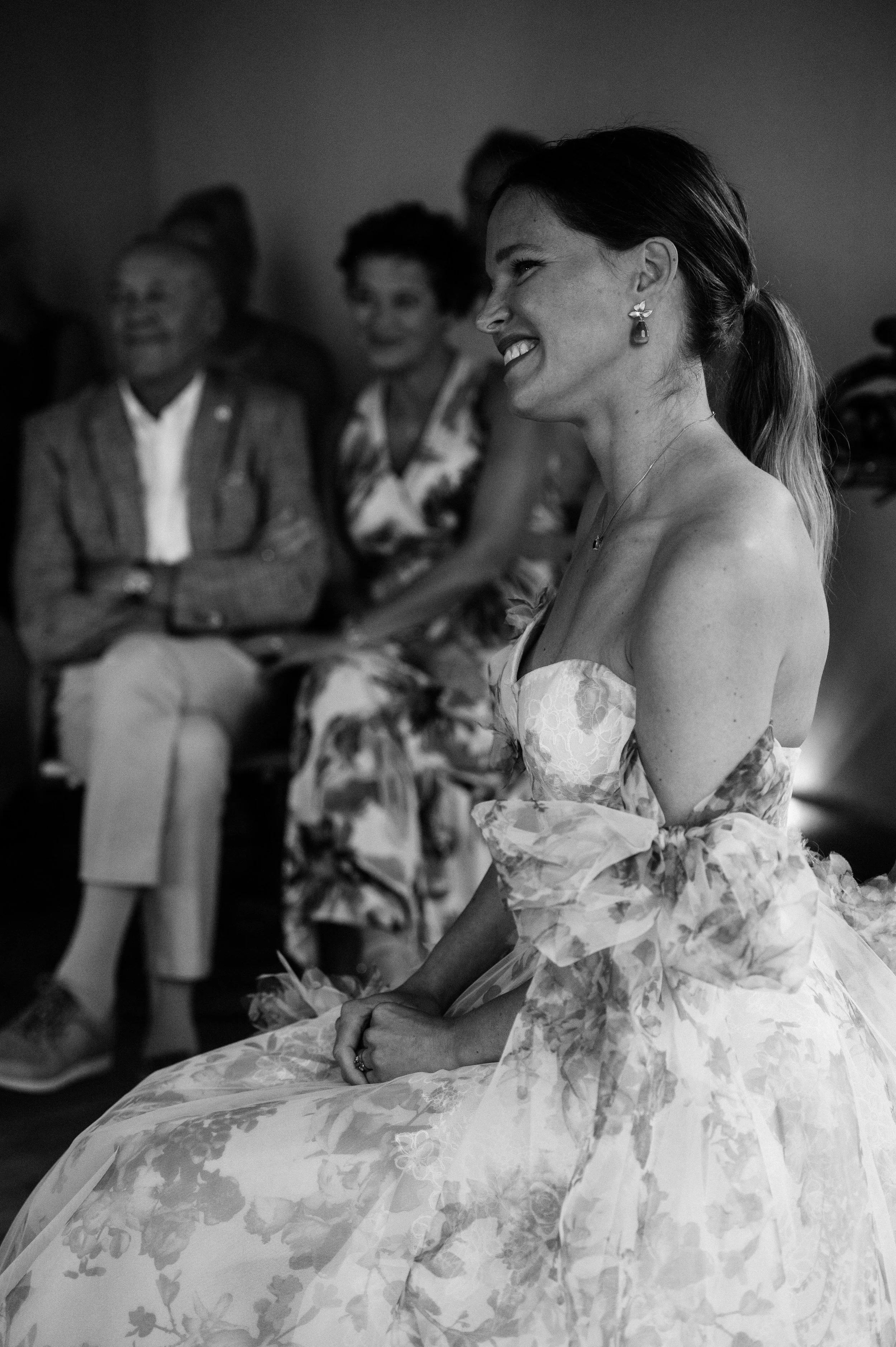 A woman in a strapless floral dress smiling during a gathering, with three women in the background also seated and smiling.