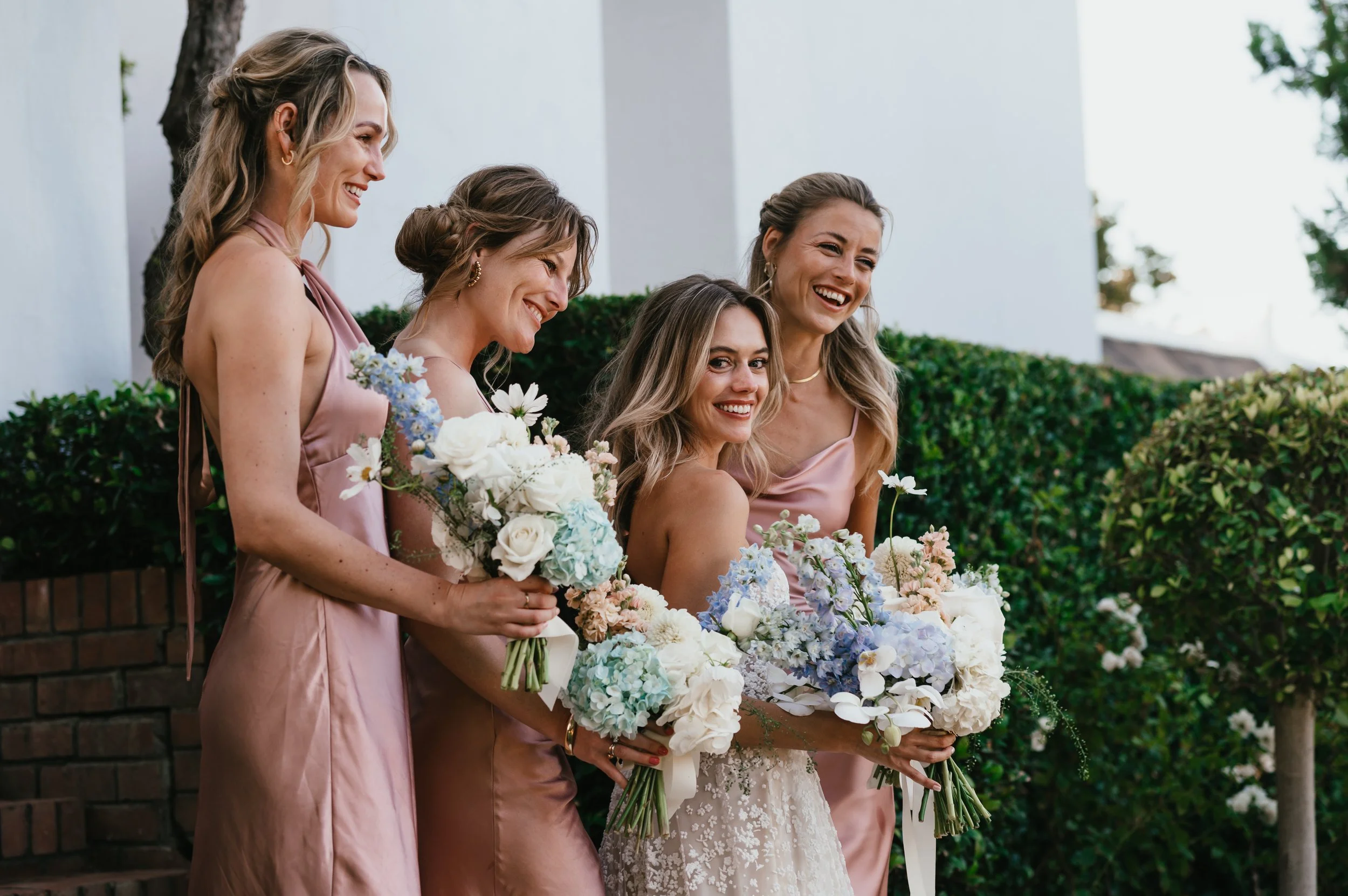 Group of five women, one in a wedding dress holding a bouquet, with four women in matching pink dresses, standing outdoors with green bushes and trees.