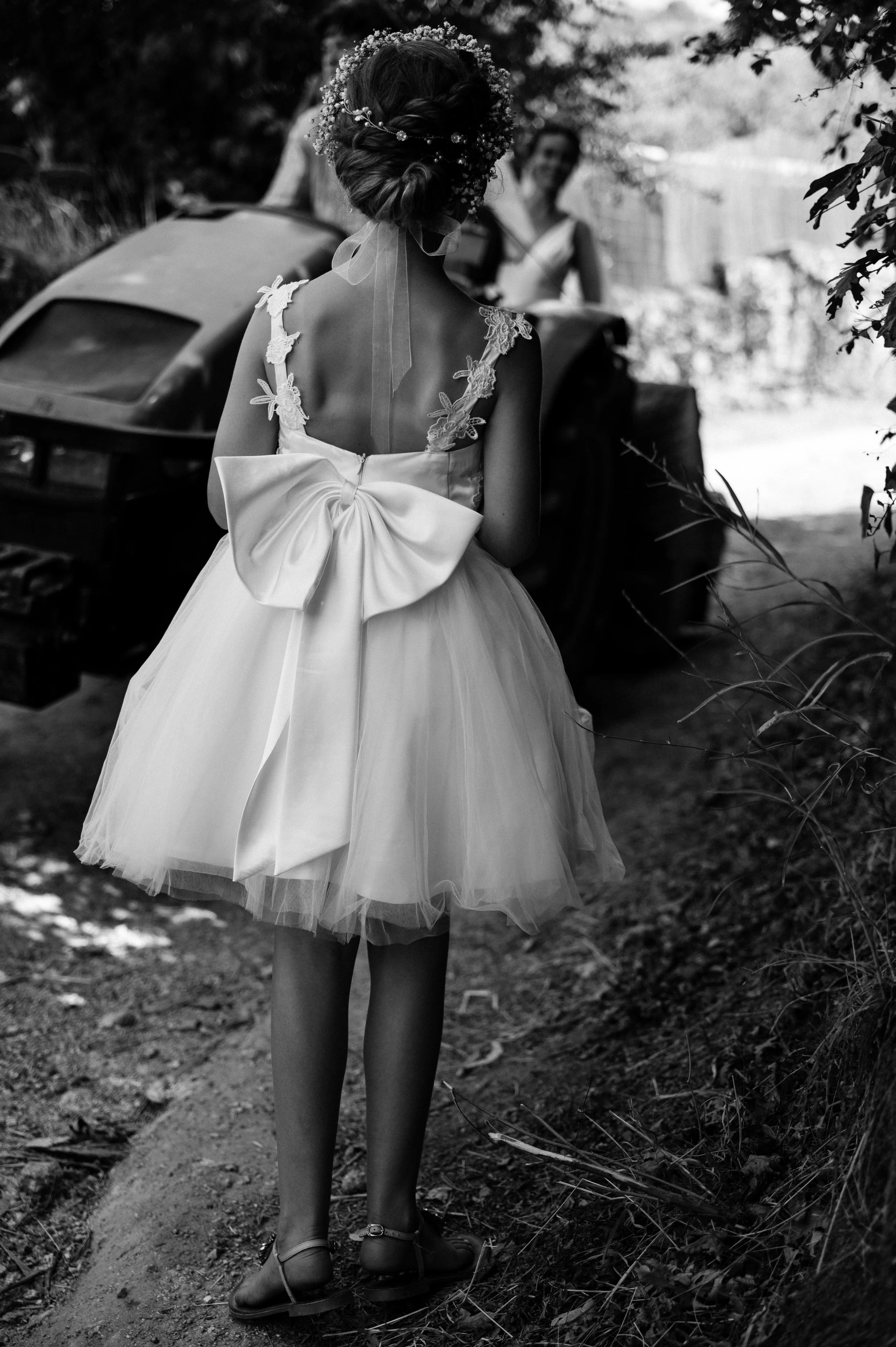 A girl in a fancy dress with a large bow on the back, surrounded by greenery, near a car, with a woman smiling in the background.