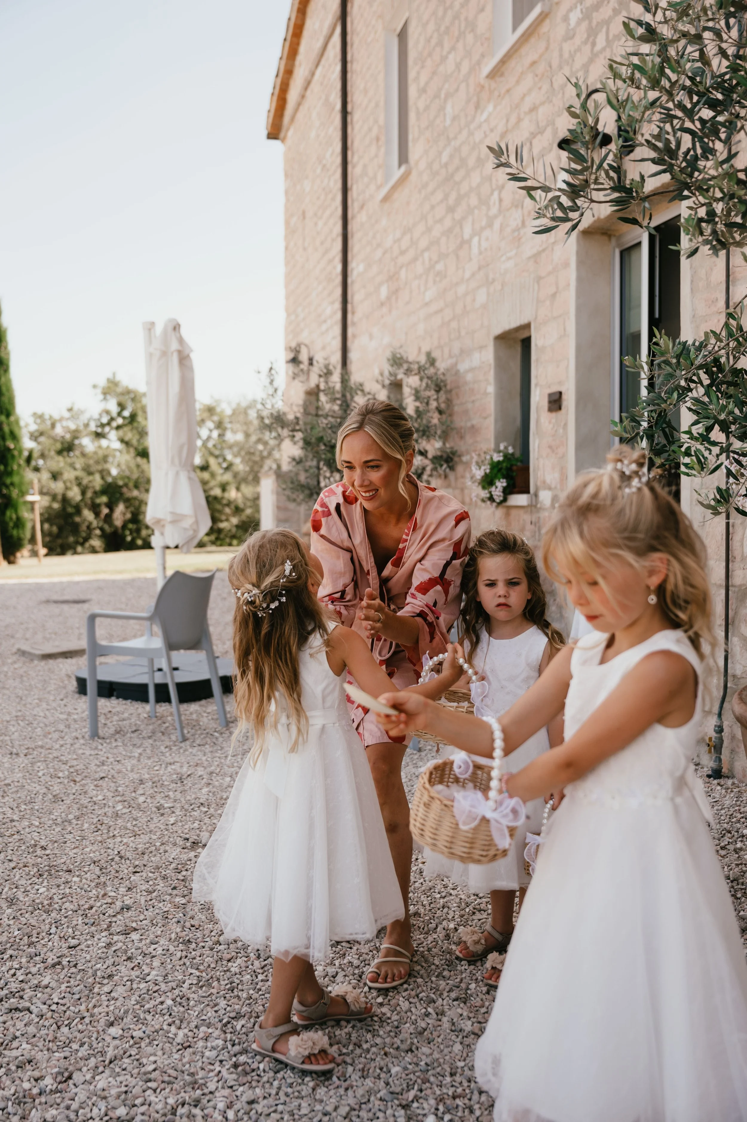 A woman and three young girls dressed in white dresses meet on a gravel outdoor space next to a stone house, with one girl holding a basket and jewelry. The woman is smiling and interacting with the girls.