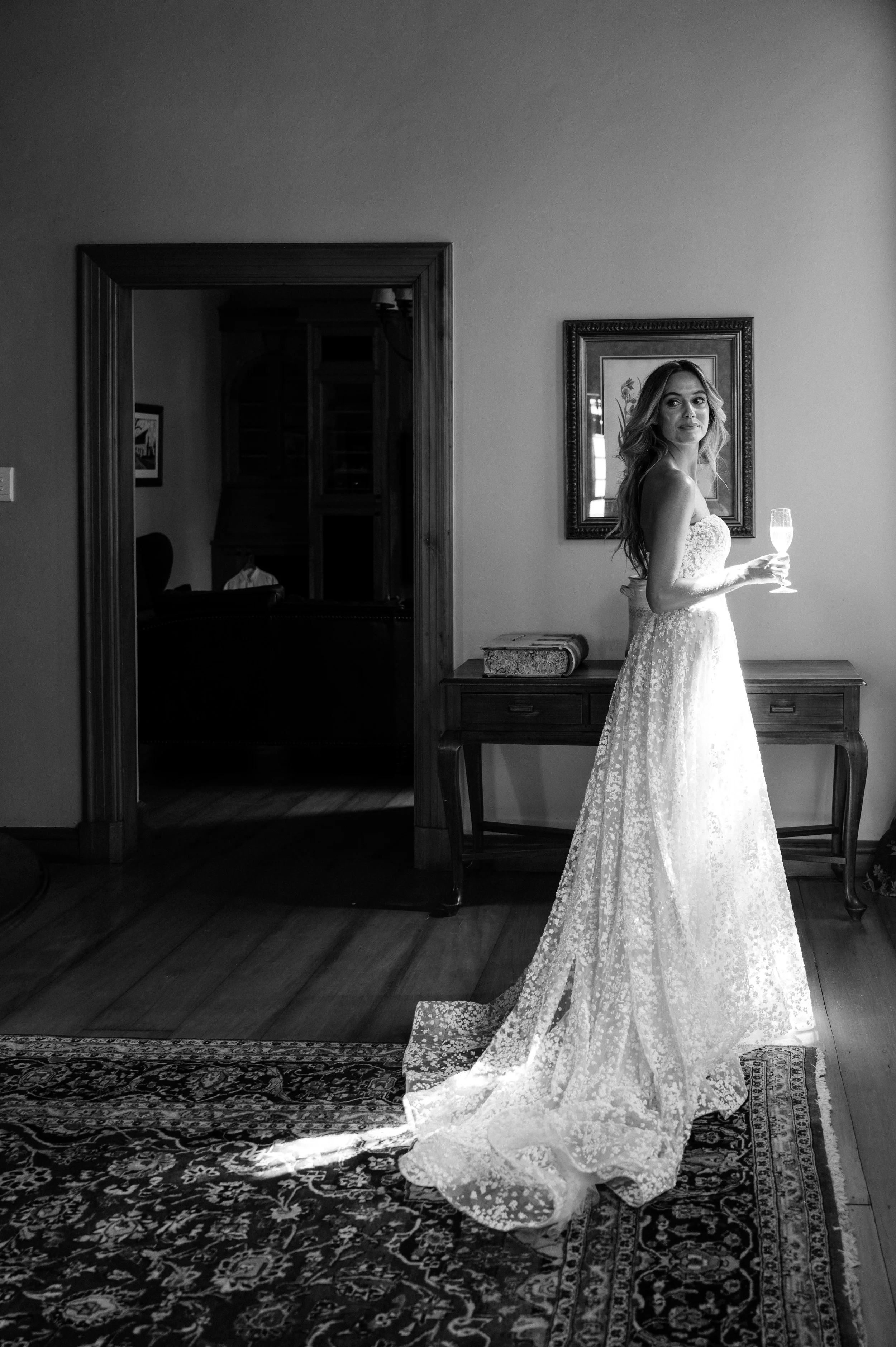 A woman in a wedding dress holding a champagne glass, standing on a rug in a room with wooden floors, a wooden table, and framed pictures on the wall in black and white photography style.