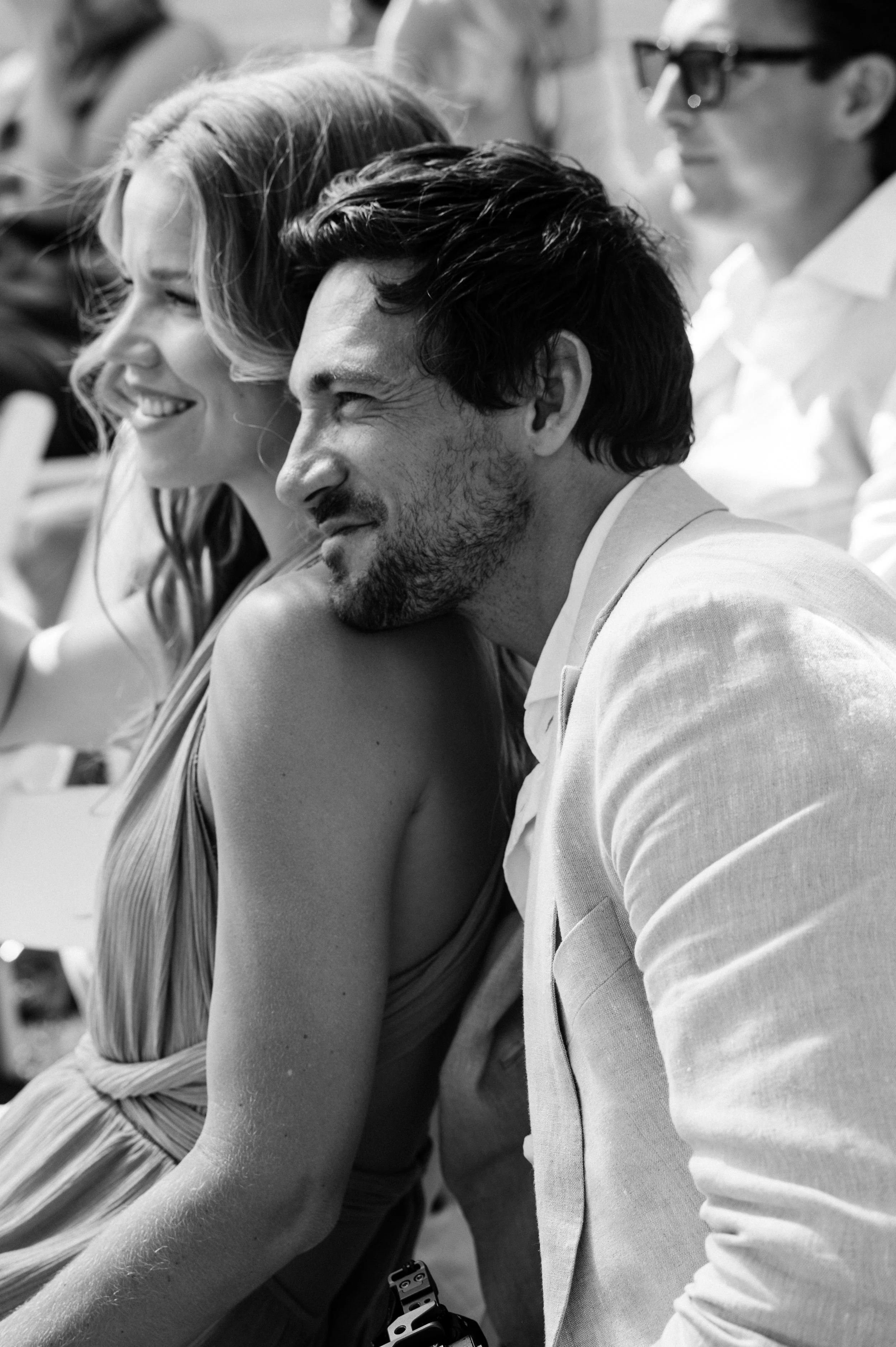 A black and white photo of a man and woman sitting closely, smiling, with the woman resting her head on the man's shoulder at an outdoor event.