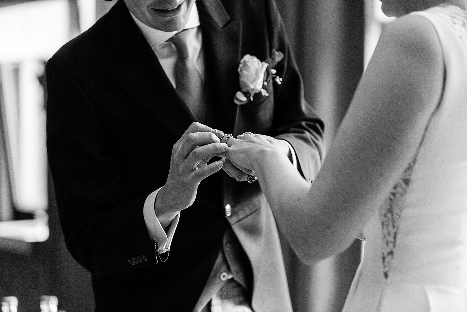 A black and white photo of a man in a suit exchanging rings with a woman in a wedding dress during a wedding ceremony.