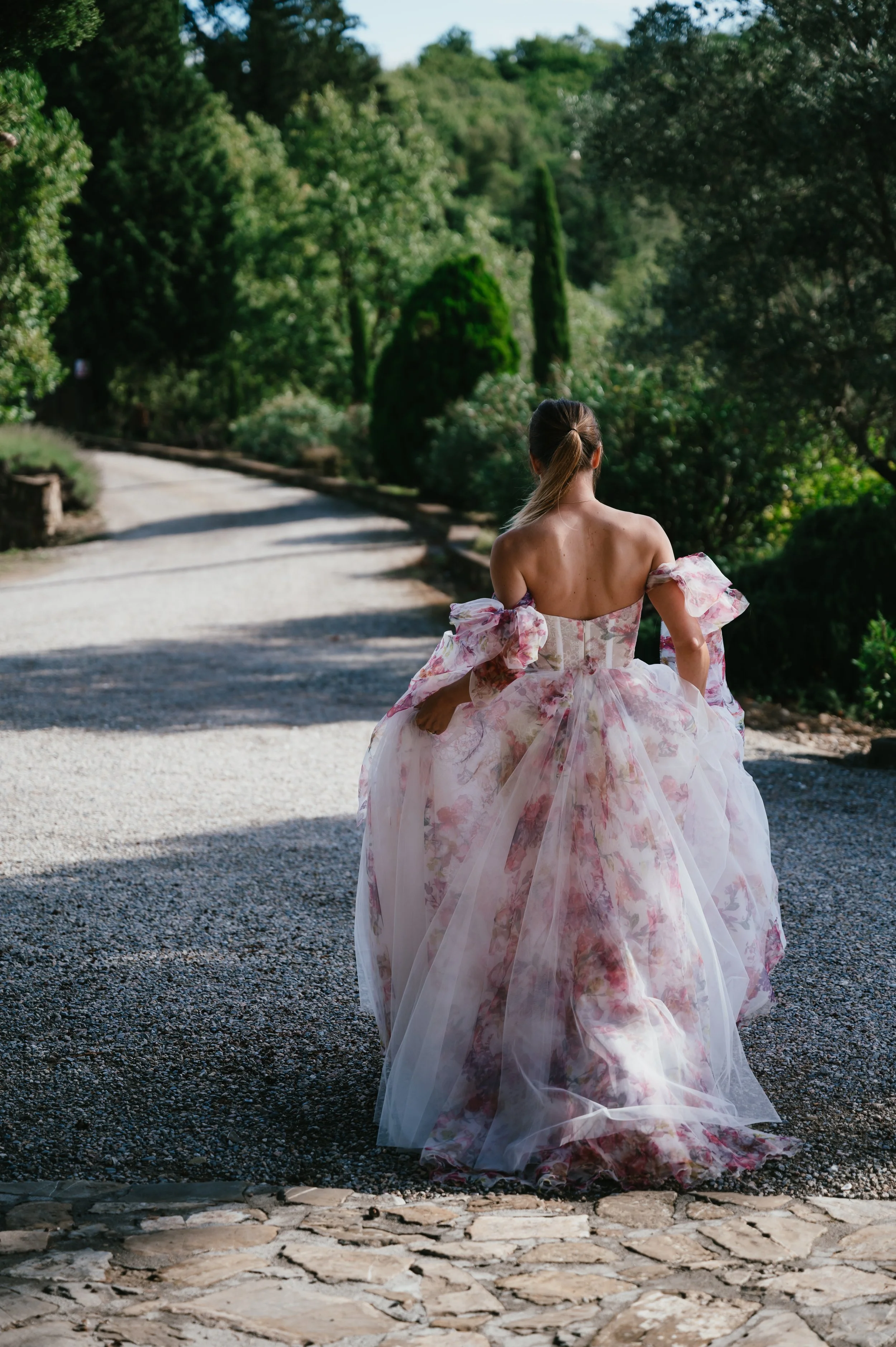 A woman in an off-the-shoulder, floral, pink ball gown walking down a gravel and stone path in a lush green park or garden.
