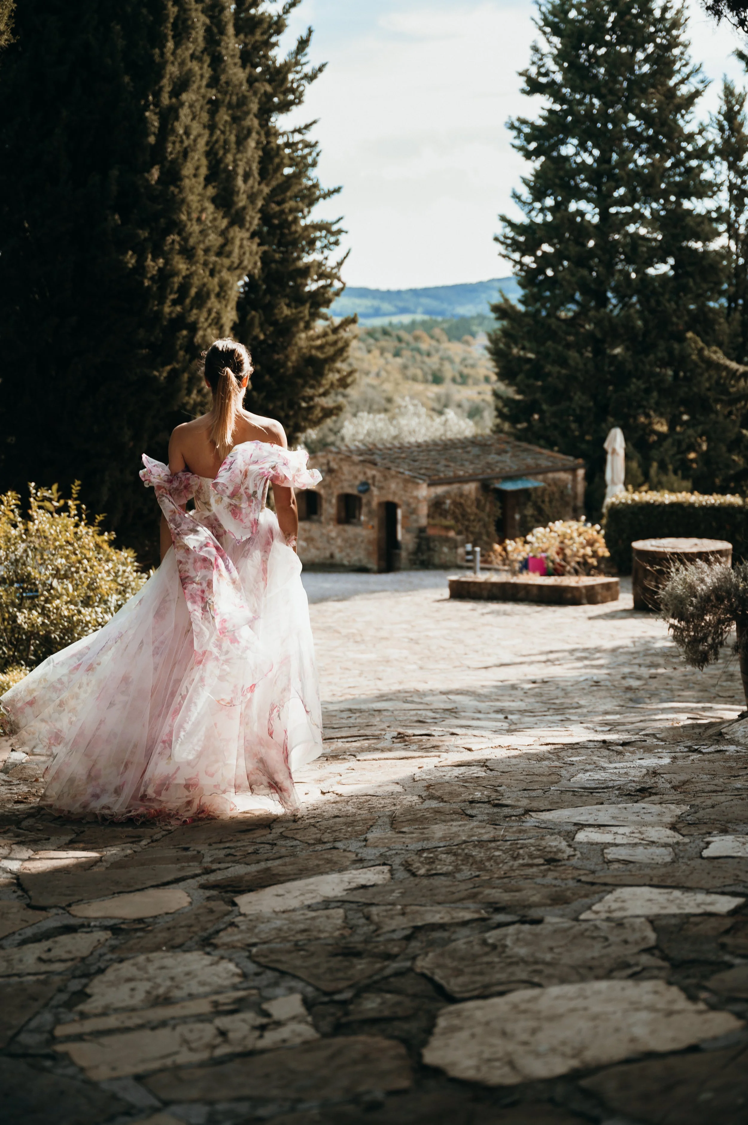 A bride in a flowing pink floral dress by Edwin Oudshoorn walking away on a stone pathway in a garden area with trees and a small stone building in the background.