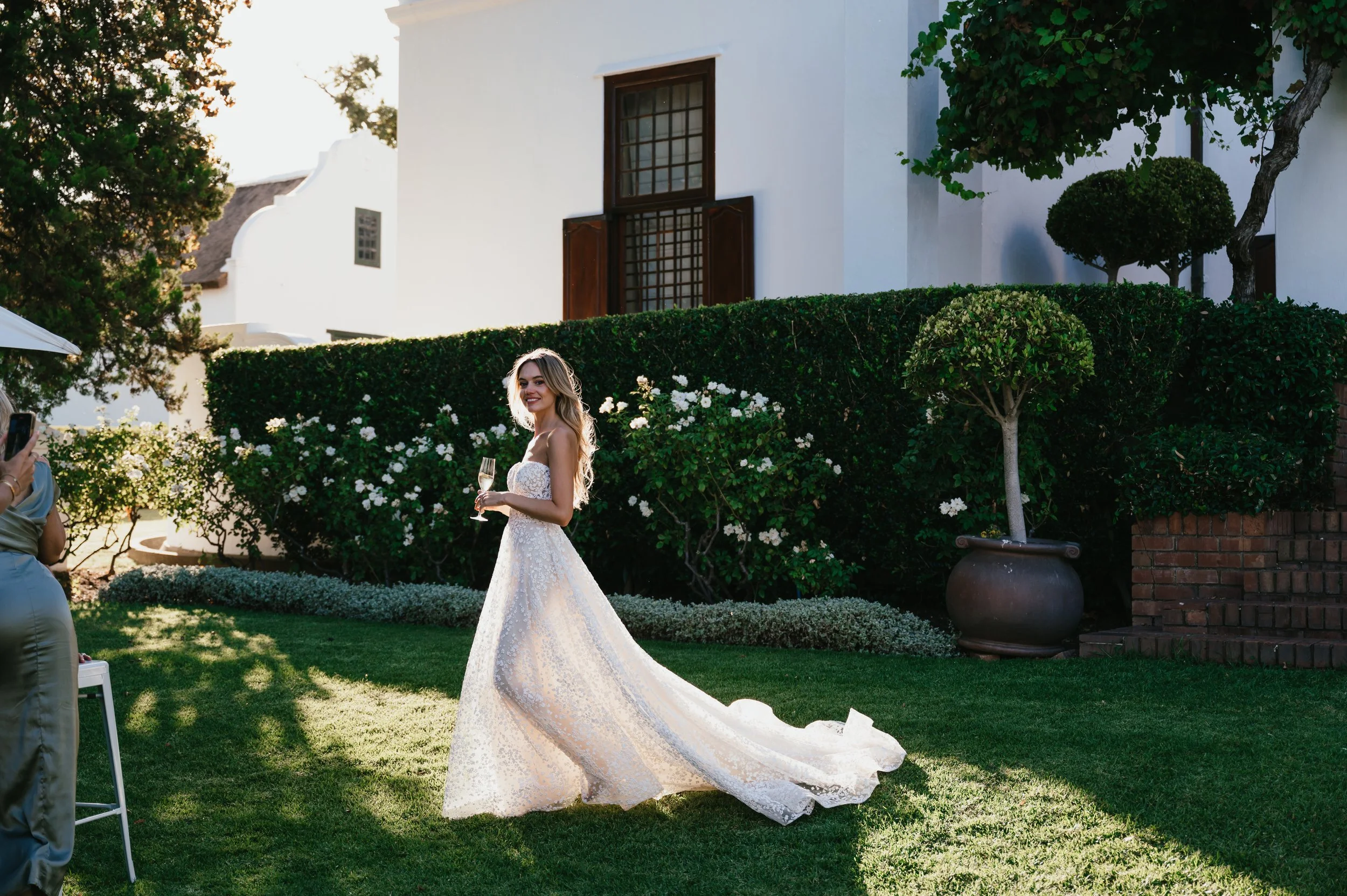 A bride in a white wedding dress holding a glass of champagne, smiling in a garden with green bushes and trees, near a white building with wooden shutters, during sunlight.