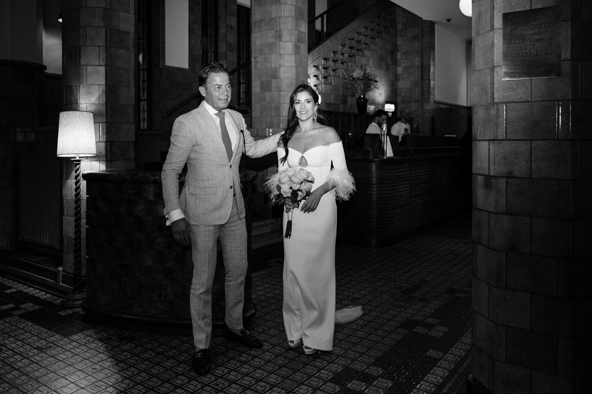 A black-and-white photo of a woman in a wedding dress holding a bouquet of flowers, standing beside a man in a suit in a hotel or restaurant lobby.